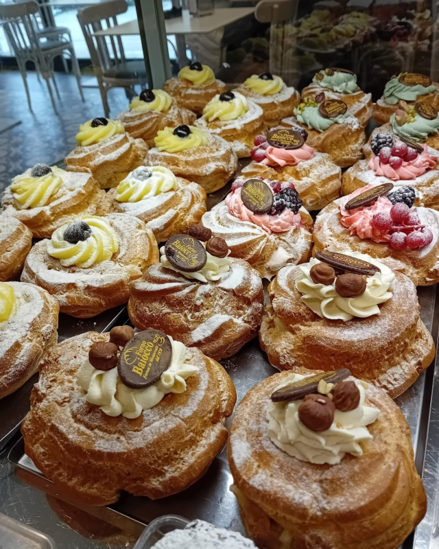Assortimento di bignè ripieni di crema, frutta e cioccolato, disposti su un vassoio di metallo in una vetrina di pasticceria.