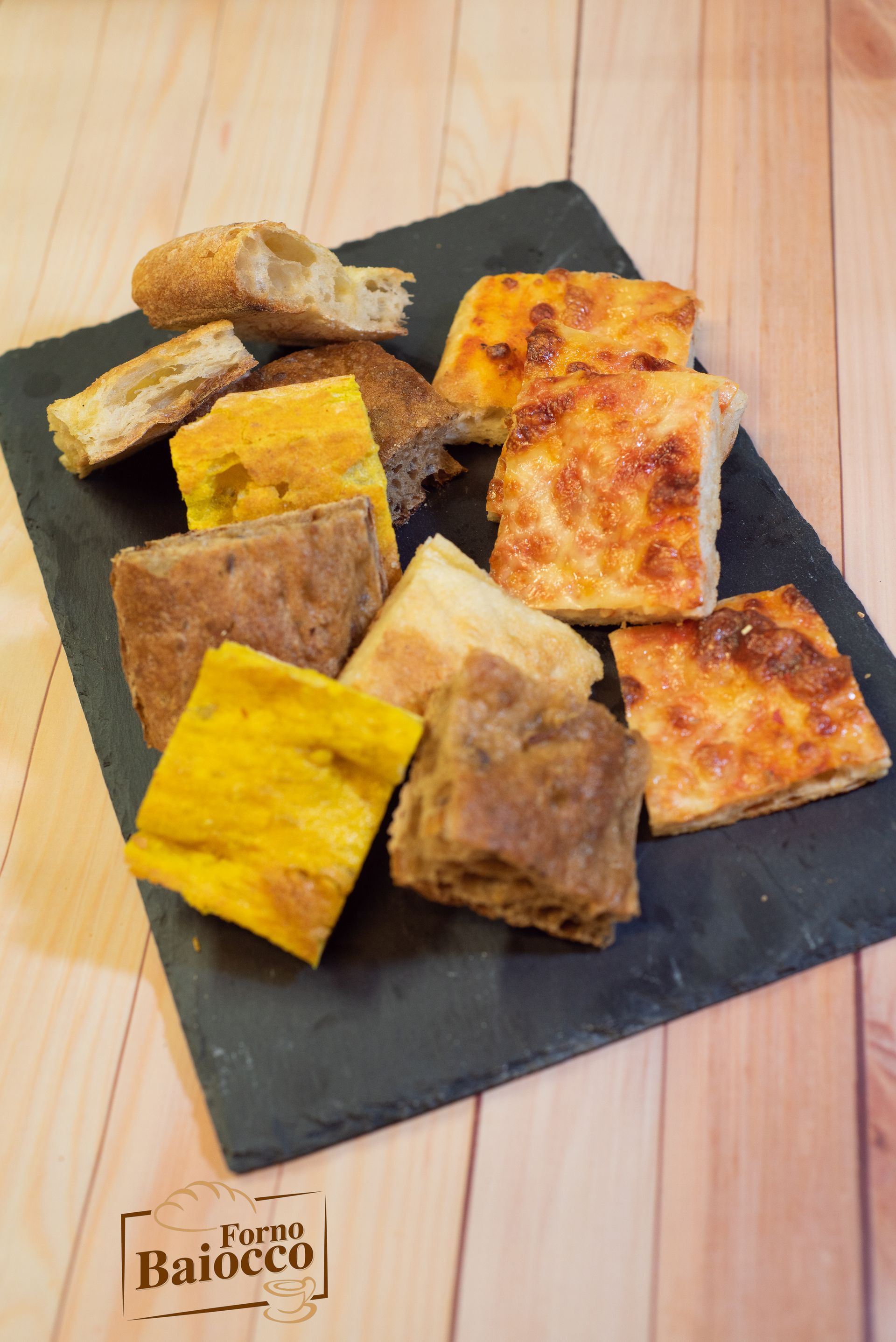 Vari pezzi quadrati di focaccia, tra cui varietà dorate e con formaggio, disposti su un vassoio di ardesia nera.