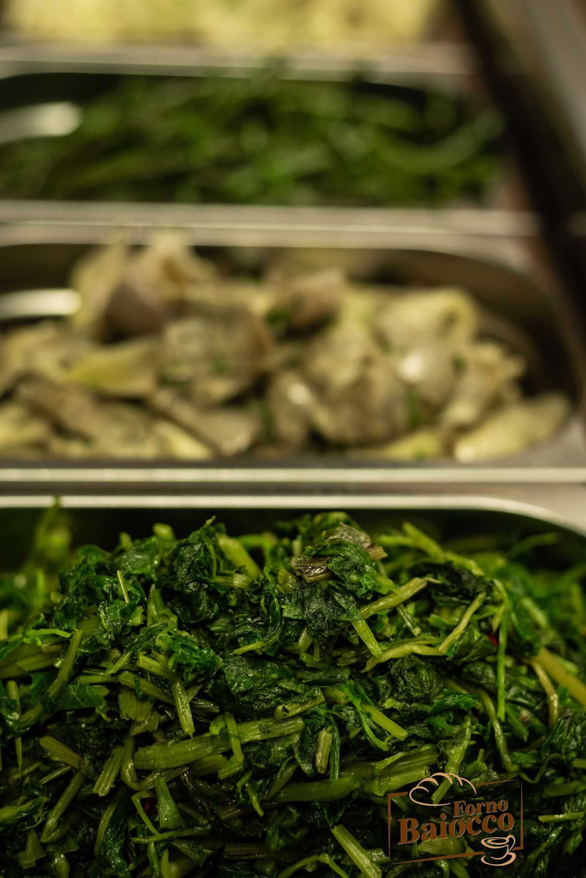 Primo piano di vassoi di cibo in un buffet contenenti verdure a foglia verde cotte, carciofi e altre verdure verdi.