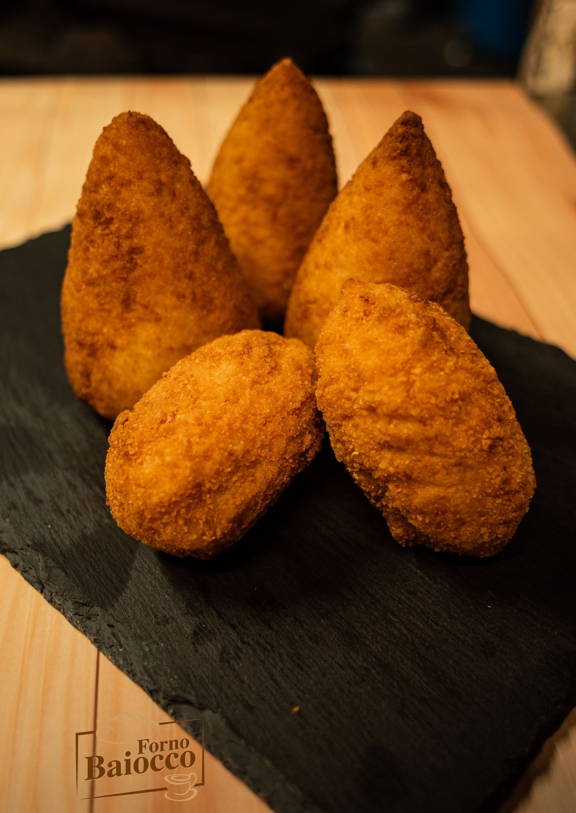 Cinque arancini fritti di colore dorato, disposti su una lastra di ardesia nera appoggiata su una superficie di legno.
