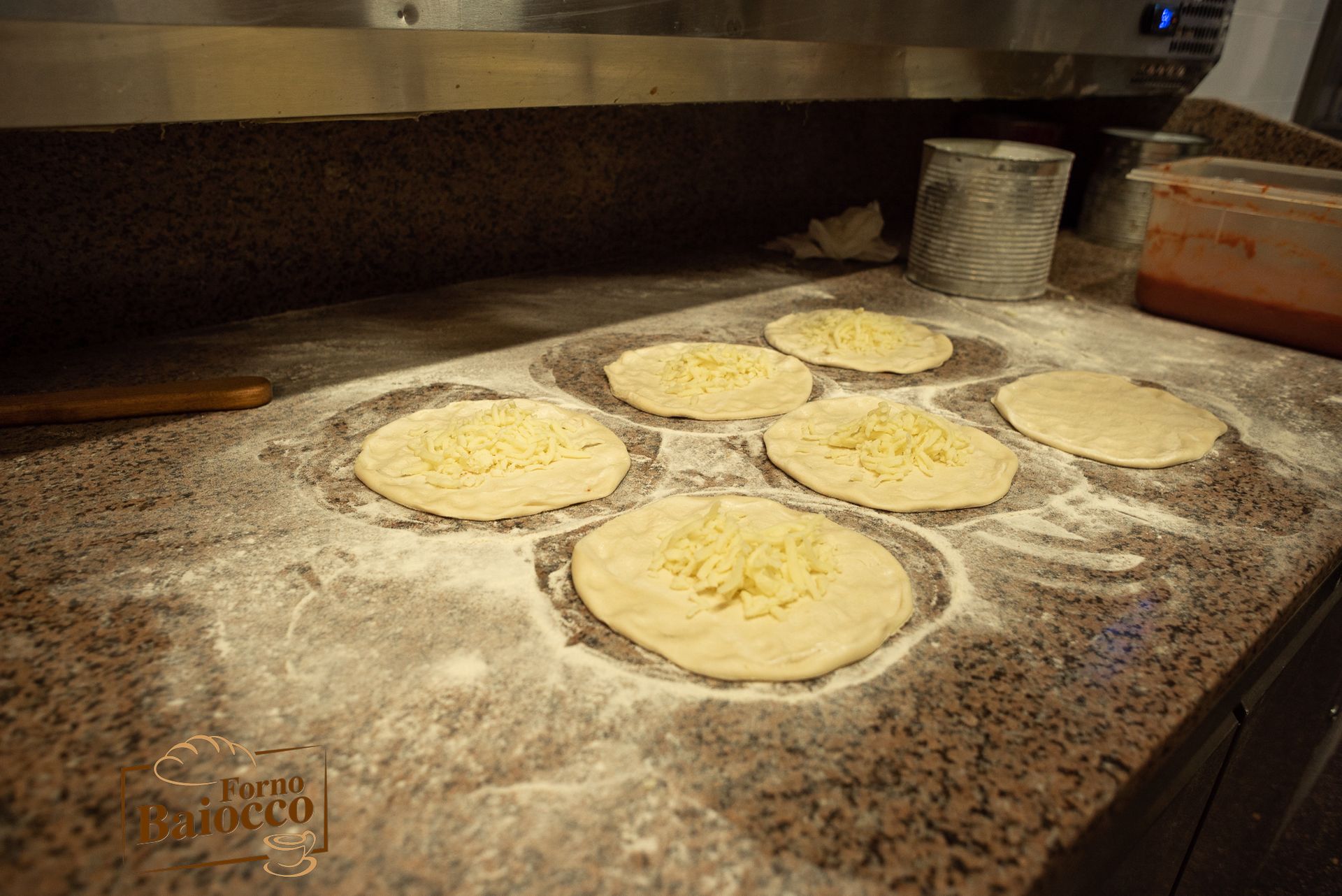 Sei dischi di pasta per pizza ricoperti di formaggio grattugiato, disposti su un piano di lavoro in marmo infarinato