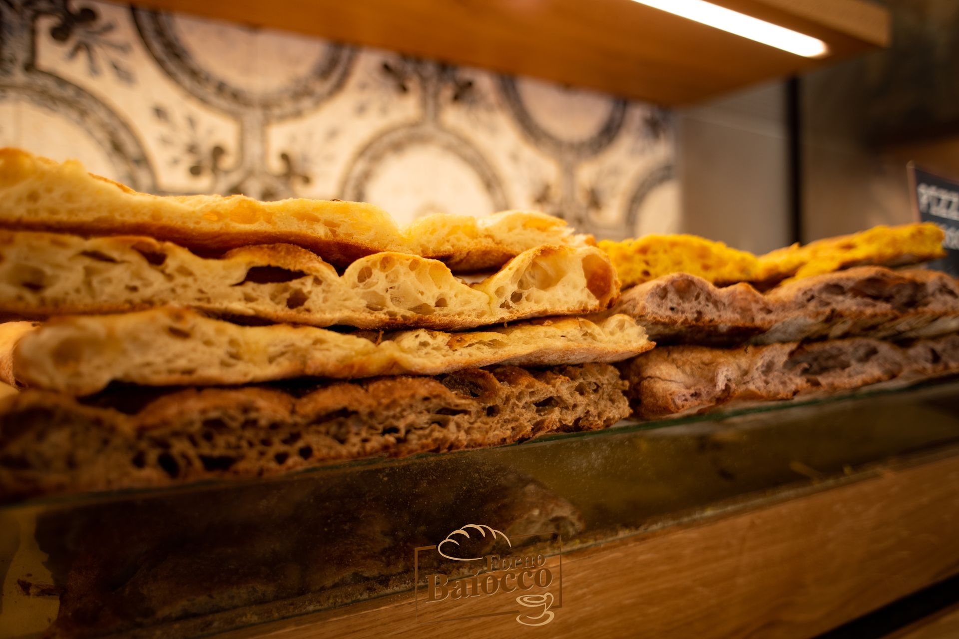 Primo piano di diverse focacce esposte su uno scaffale in una panetteria, con una parete piastrellata