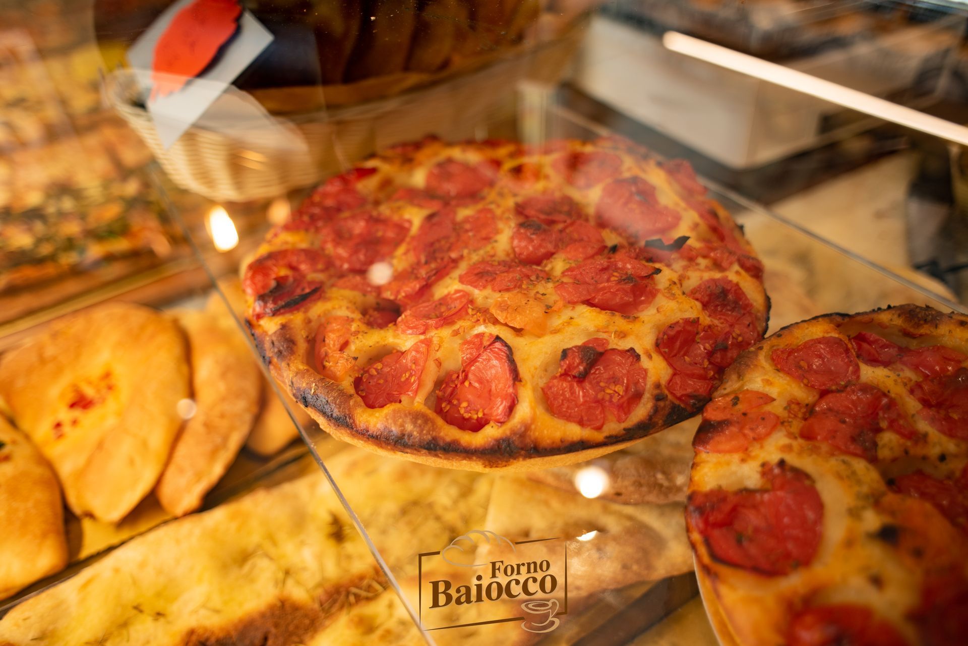 Da Forno Baiocco, la focaccia al pomodoro appena sfornata è esposta in una vetrina, insieme ad altri prodotti da forno.