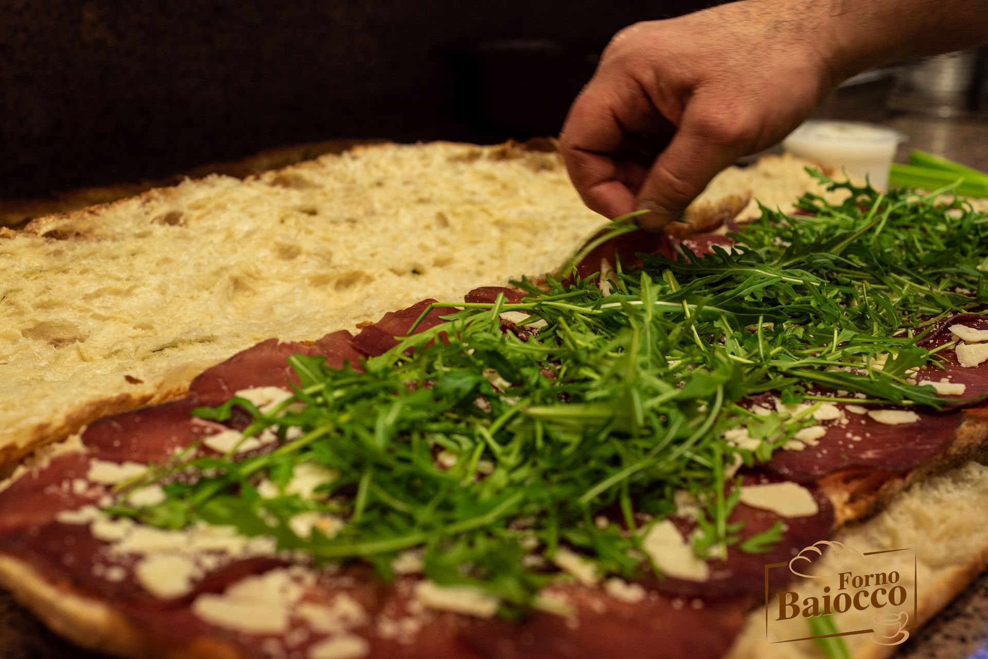 Una mano dispone la rucola sopra la bresaola e il formaggio grattugiato su un panino aperto a base di pane piatto.