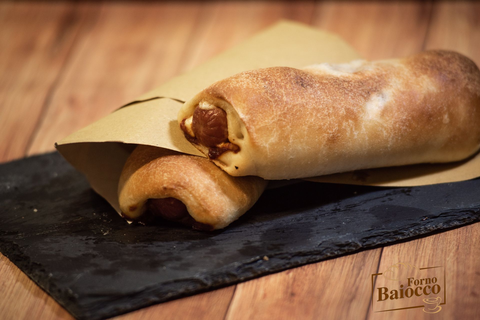 Due panini al forno ripieni di salsiccia, avvolti in carta marrone e serviti su un vassoio di ardesia nera