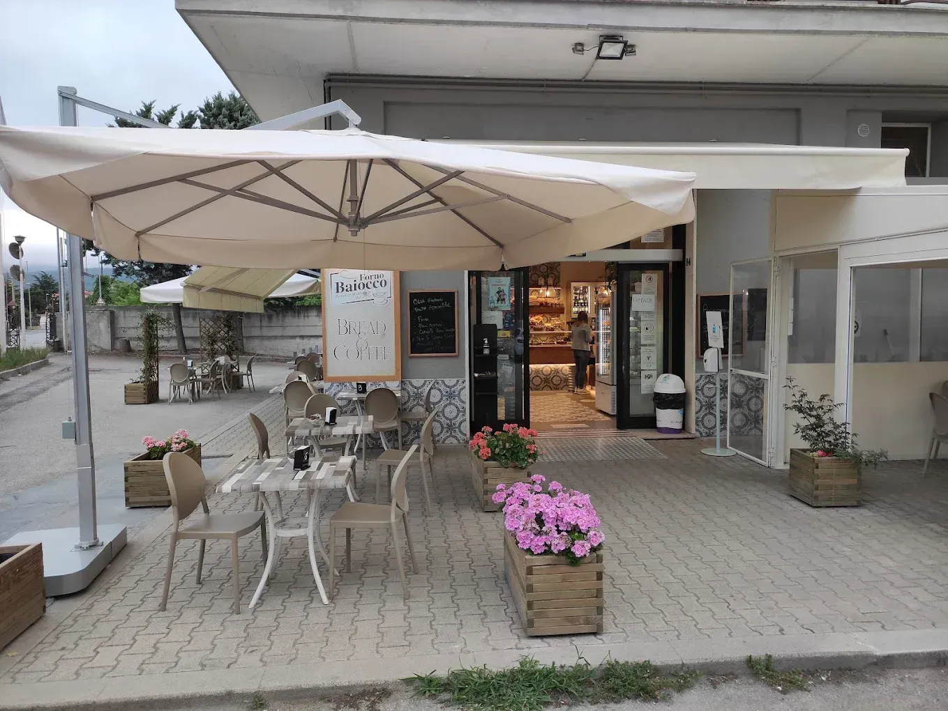 Tavolini e sedie all'aperto in un caffè, con fiori rosa in vasi di legno e un grande ombrellone beige.