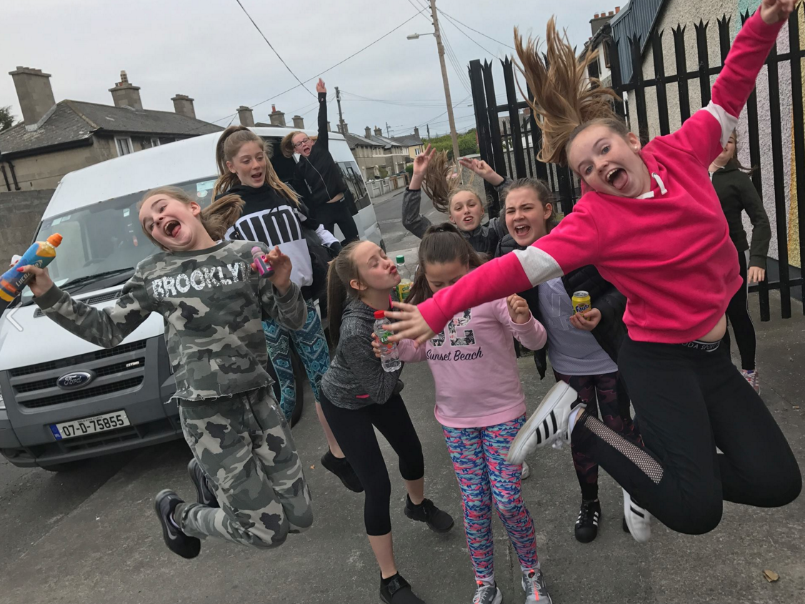 Group of girls jumping excitedly near a van, wearing casual clothes, with bright expressions.