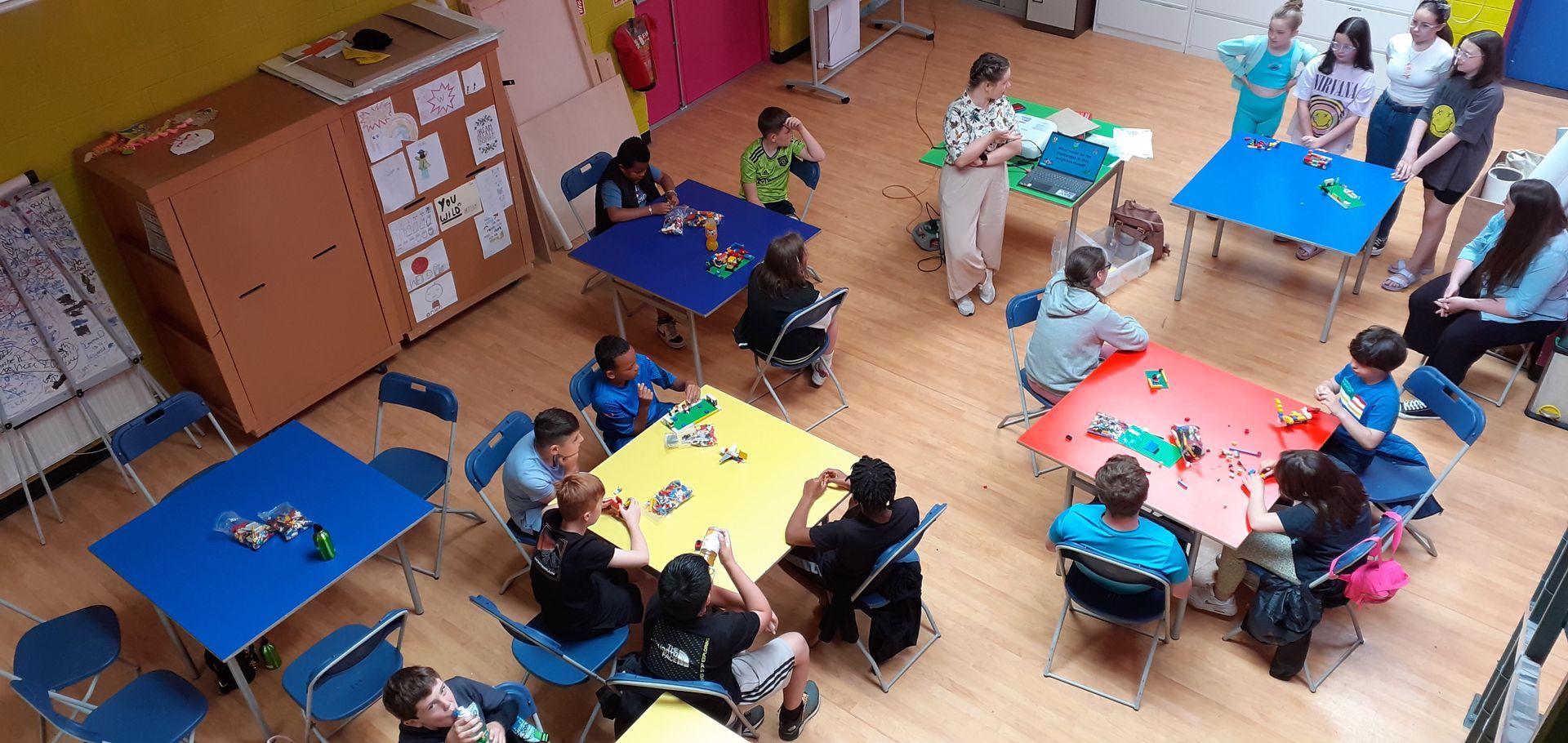 Children at colorful tables in a classroom; adults supervising.