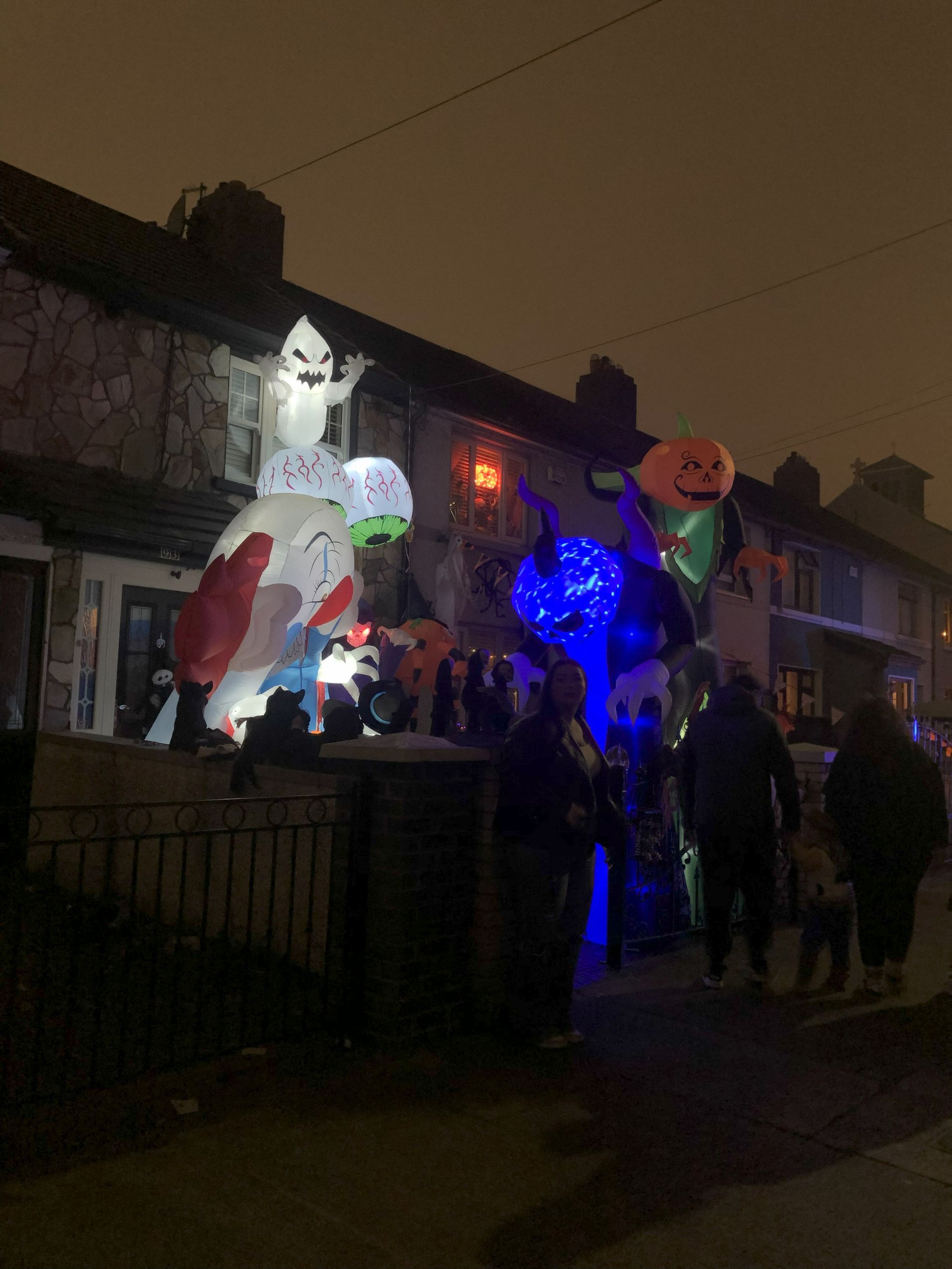 Halloween decorations on a house at night, including inflatable characters and people looking.