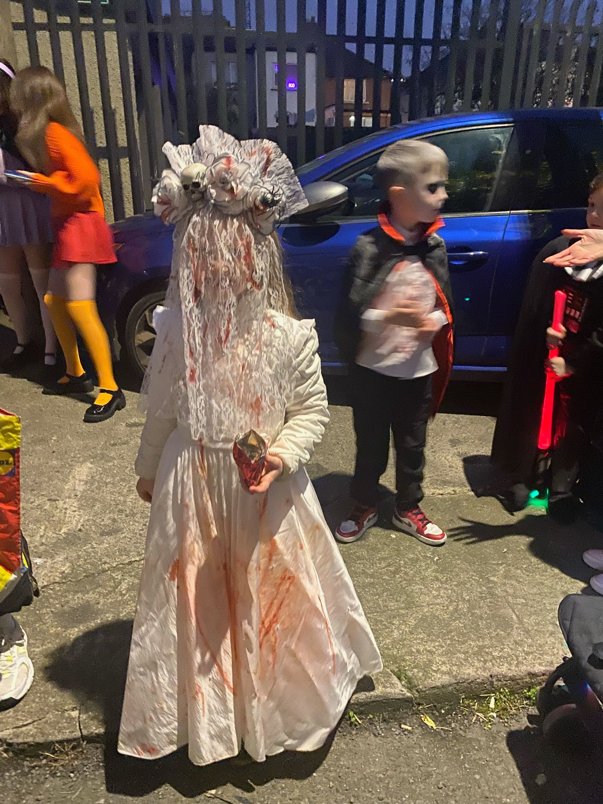 Child in a blood-splattered wedding dress costume with veil, holding a small object. Other costumed children nearby.