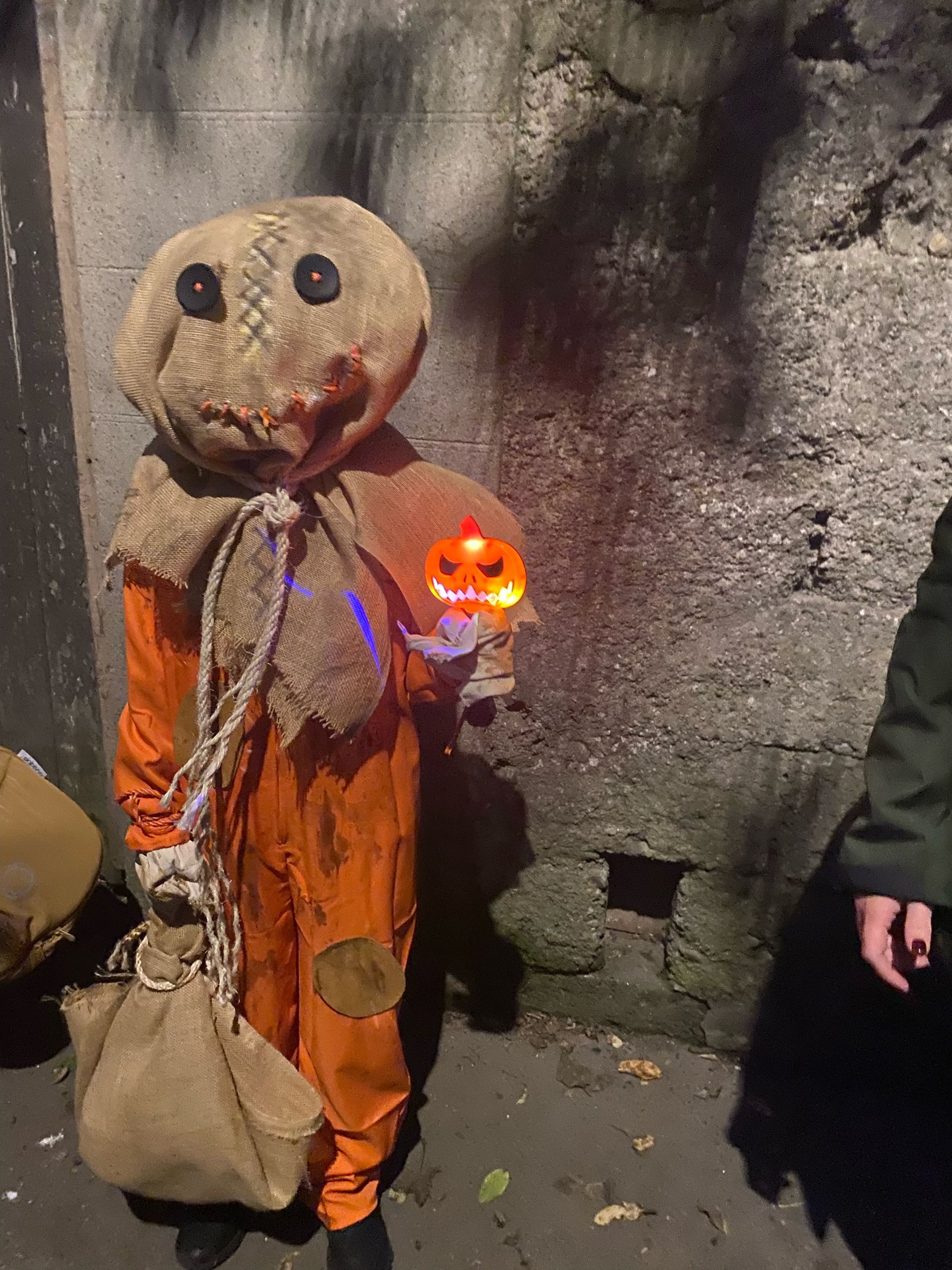 Person in  Halloween , orange jumpsuit, burlap mask, holds glowing jack-o'-lantern, stands by concrete wall, carrying bag.