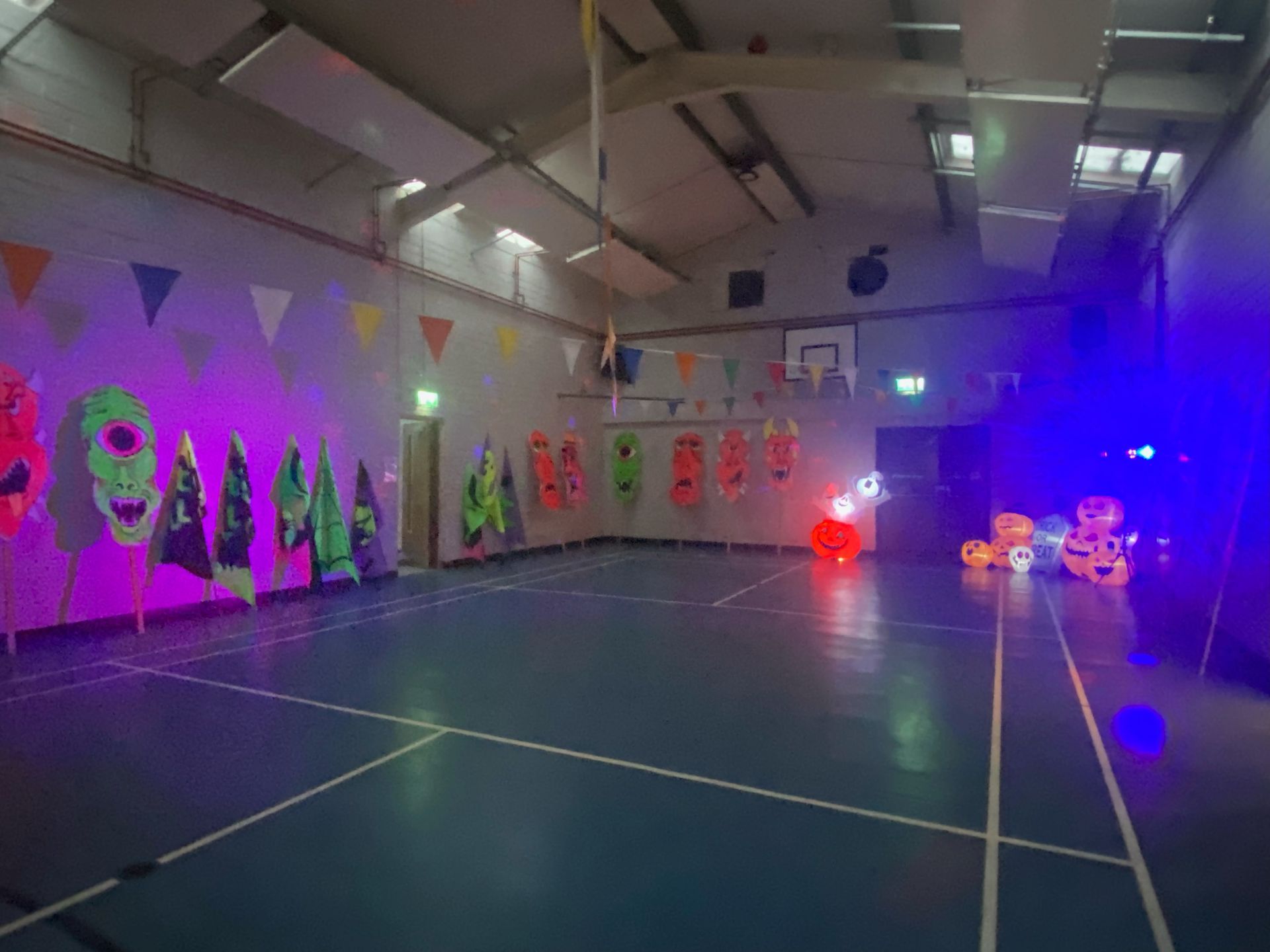 Empty gym decorated with glow-in-the-dark Halloween decorations under purple and blue lights; pennant flags hang overhead.