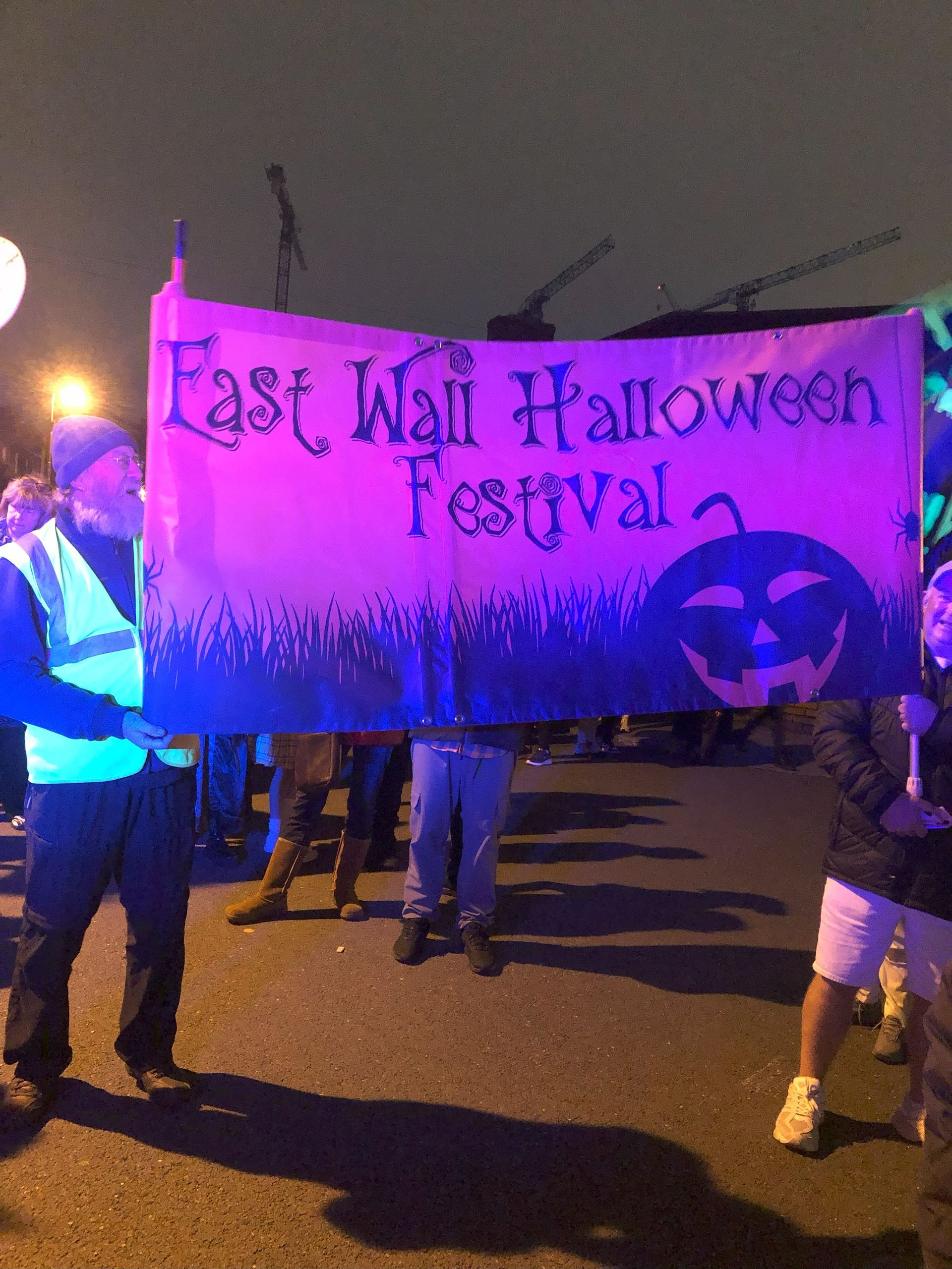 People holding a banner for the East Wall Halloween Festival, illuminated by purple light at night.