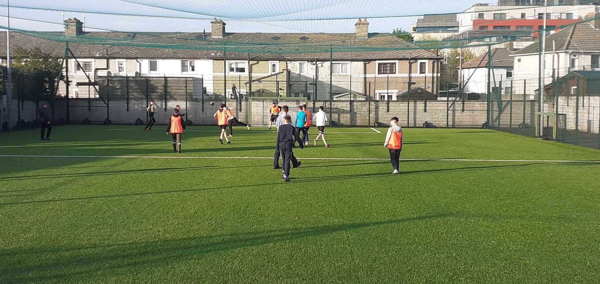 People playing on a green field surrounded by fencing and buildings.