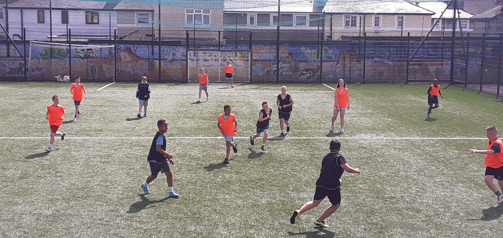 People in orange and dark shirts playing a game on a green field. Buildings in background.