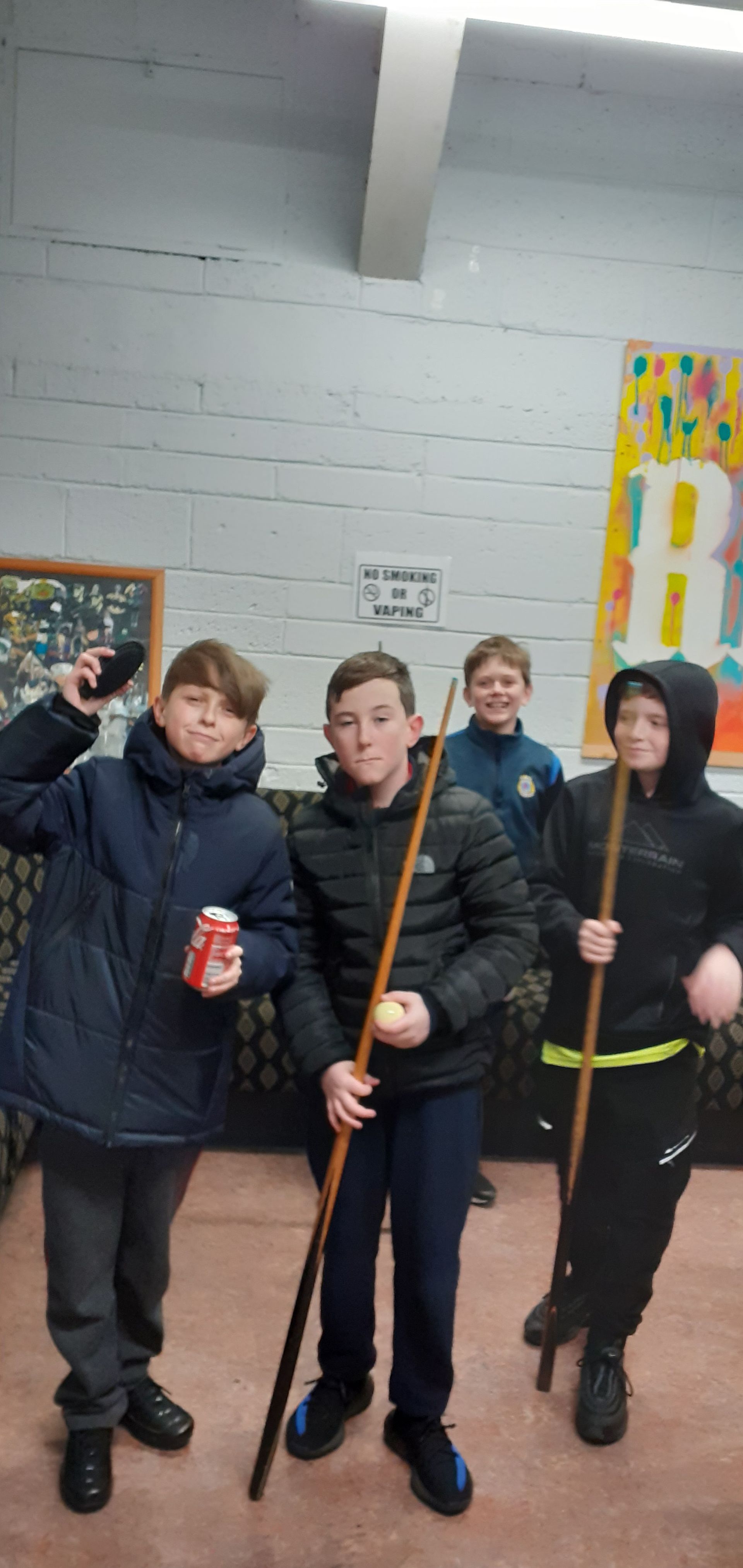 Four young people pose with pool cues and a drink. They are inside a building with a mural in the background.
