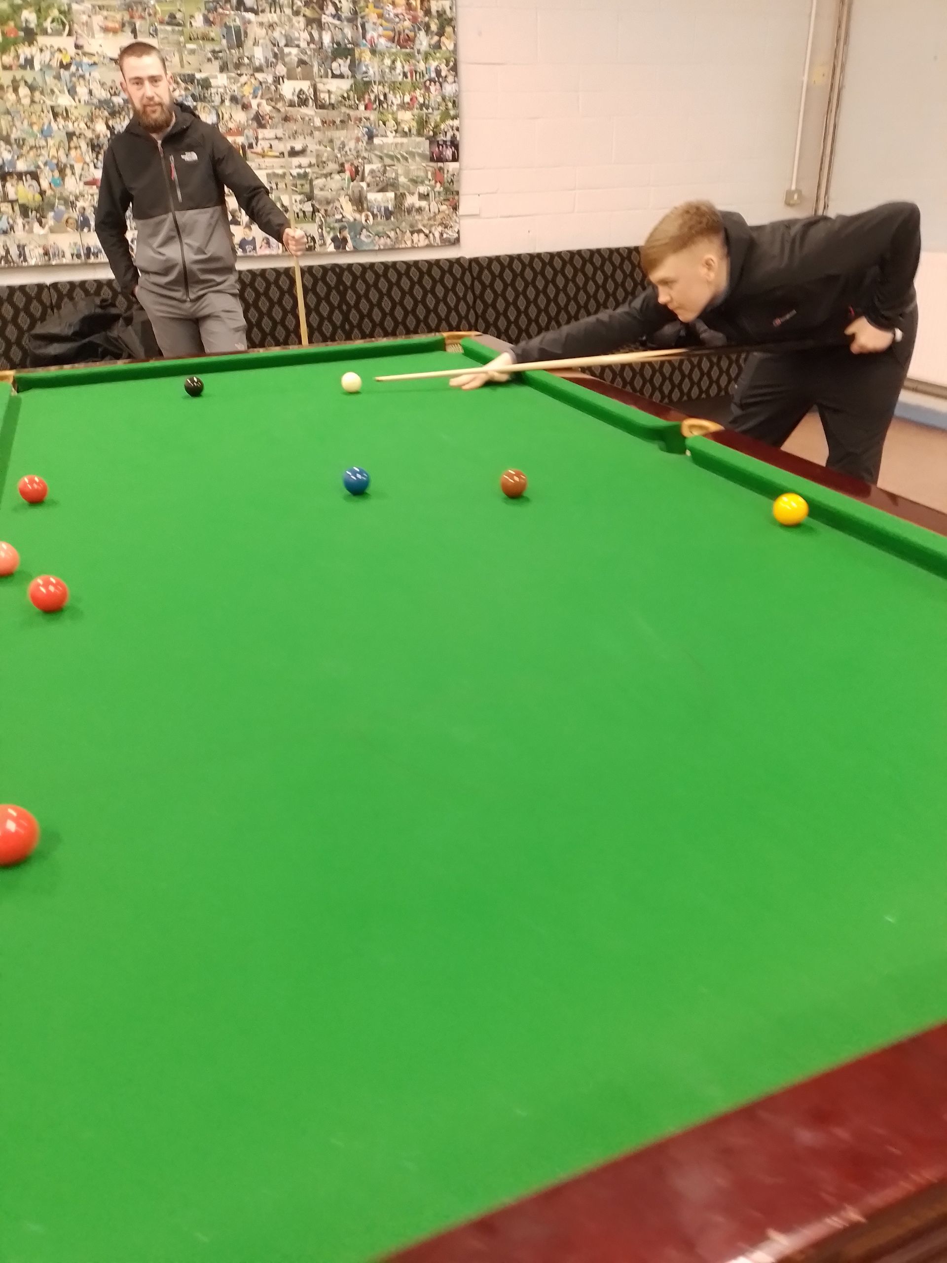 Two men playing snooker at a table. One is about to take a shot, the other watches. Green felt, balls scattered.