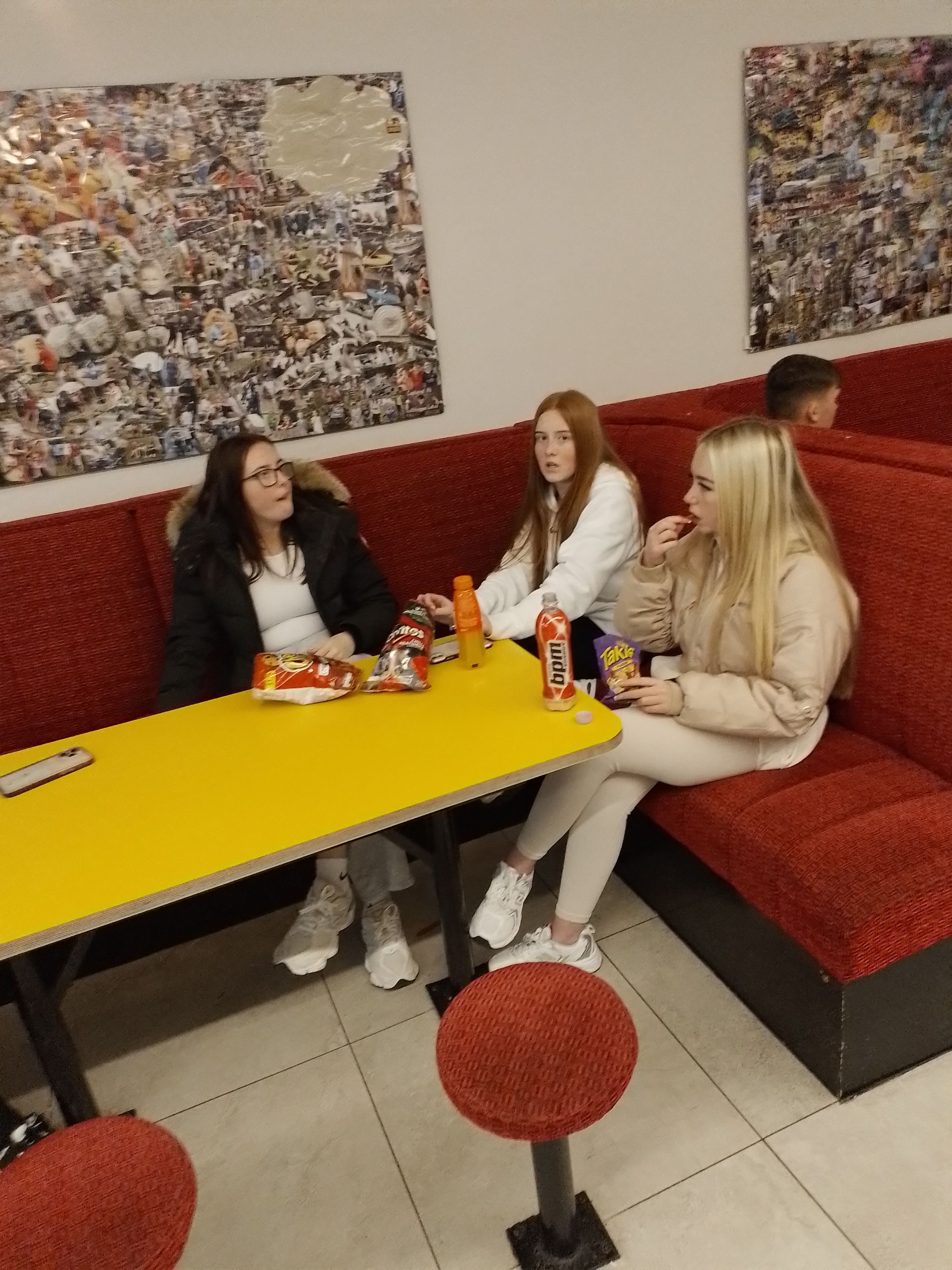 Three people seated at a yellow table in a booth, eating and talking. Red seating and a photo collage on the wall.