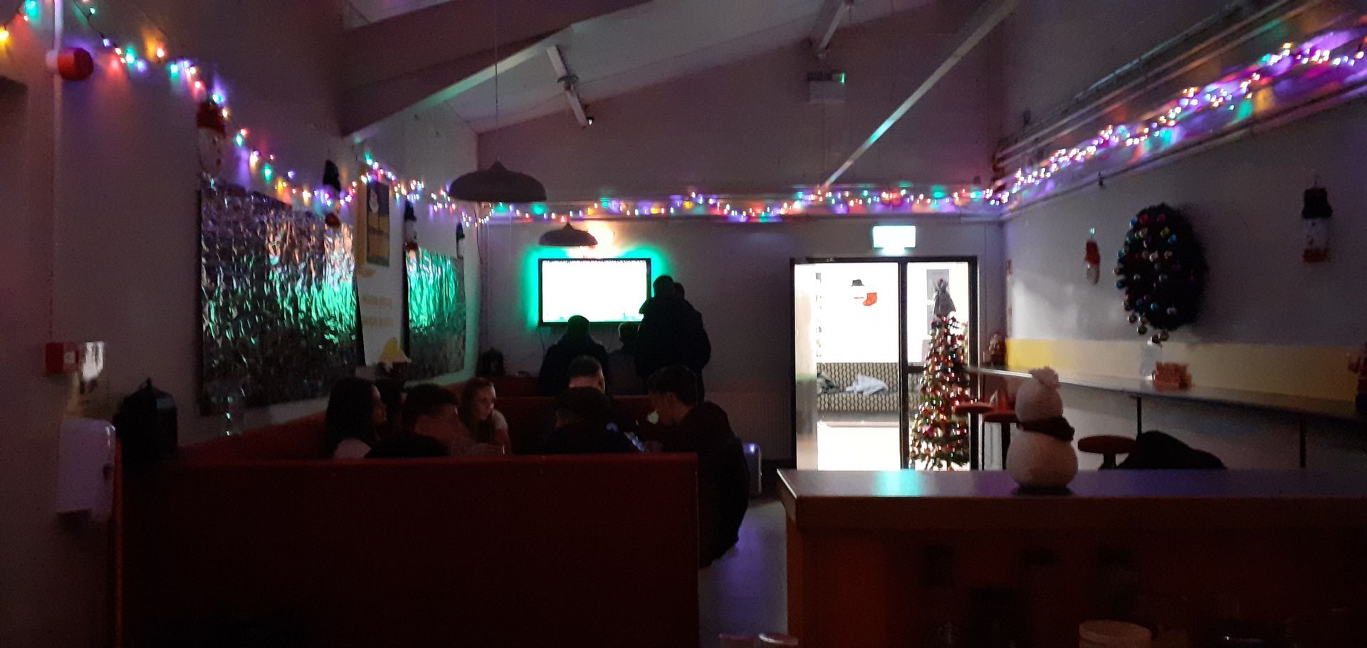 Interior shot of a room with people, Christmas decorations, and a lit tree. A TV screen is visible.