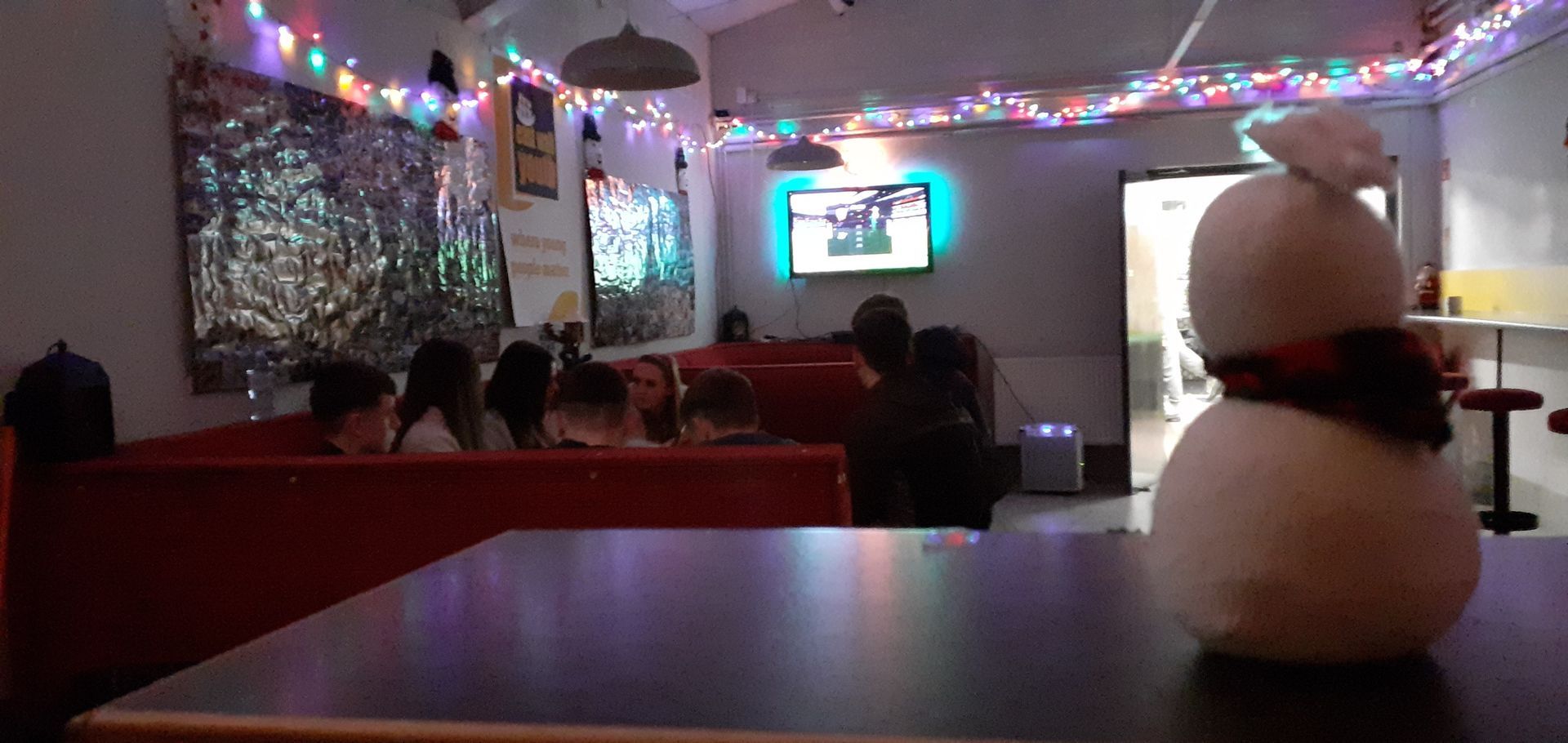 A snowman with a scarf sits on a table in a bar with Christmas lights and people in booths.