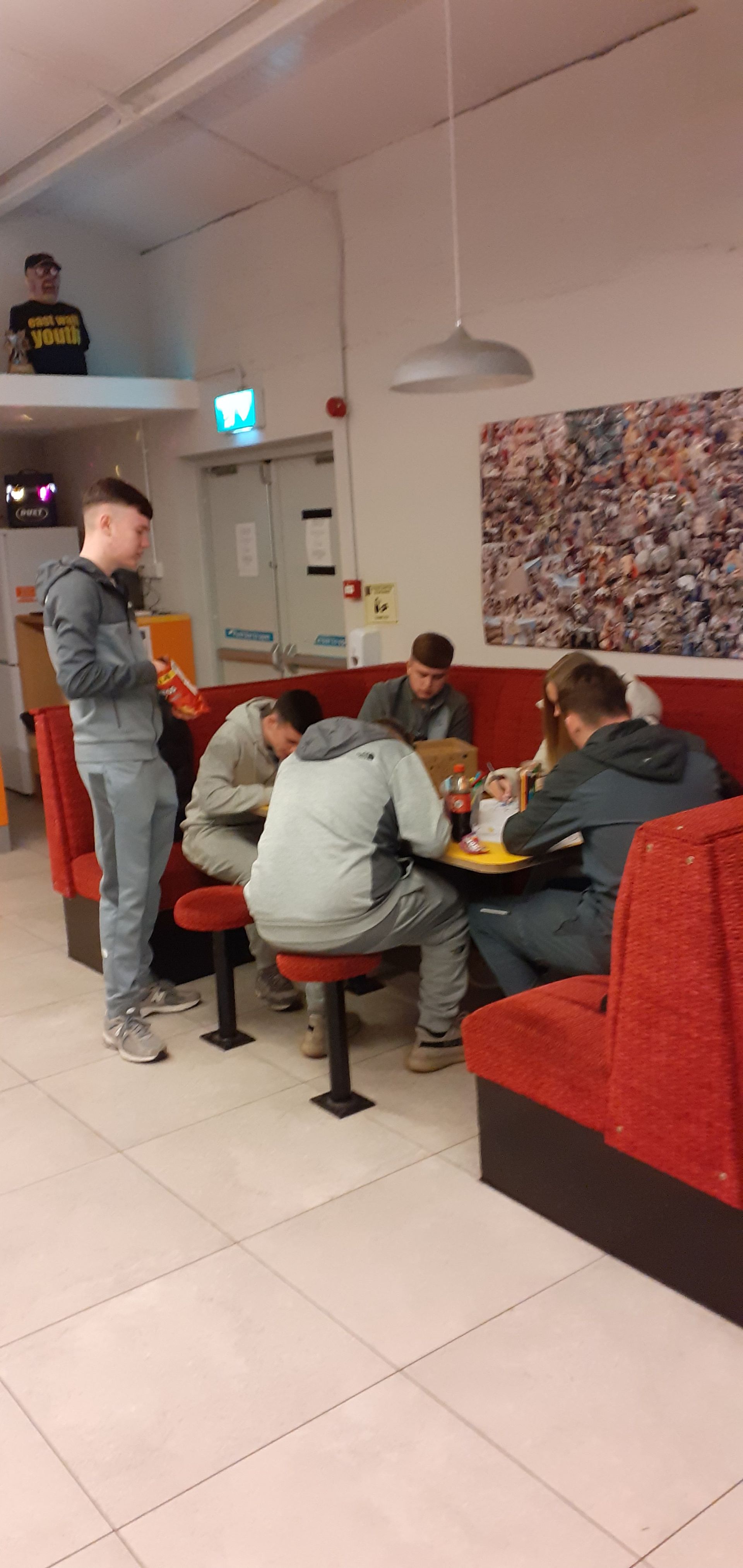 People sitting at a table in a diner. One person stands, holding something. A person is sitting on a high perch.