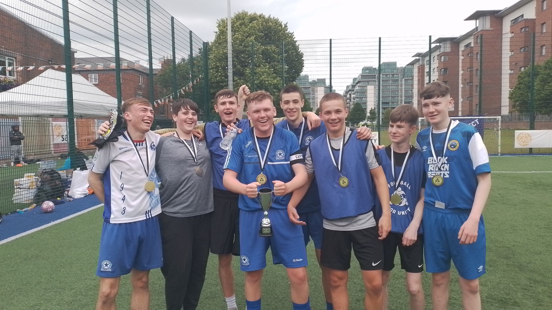 Soccer team celebrates victory, wearing blue uniforms, medals, posing on field with buildings in background.