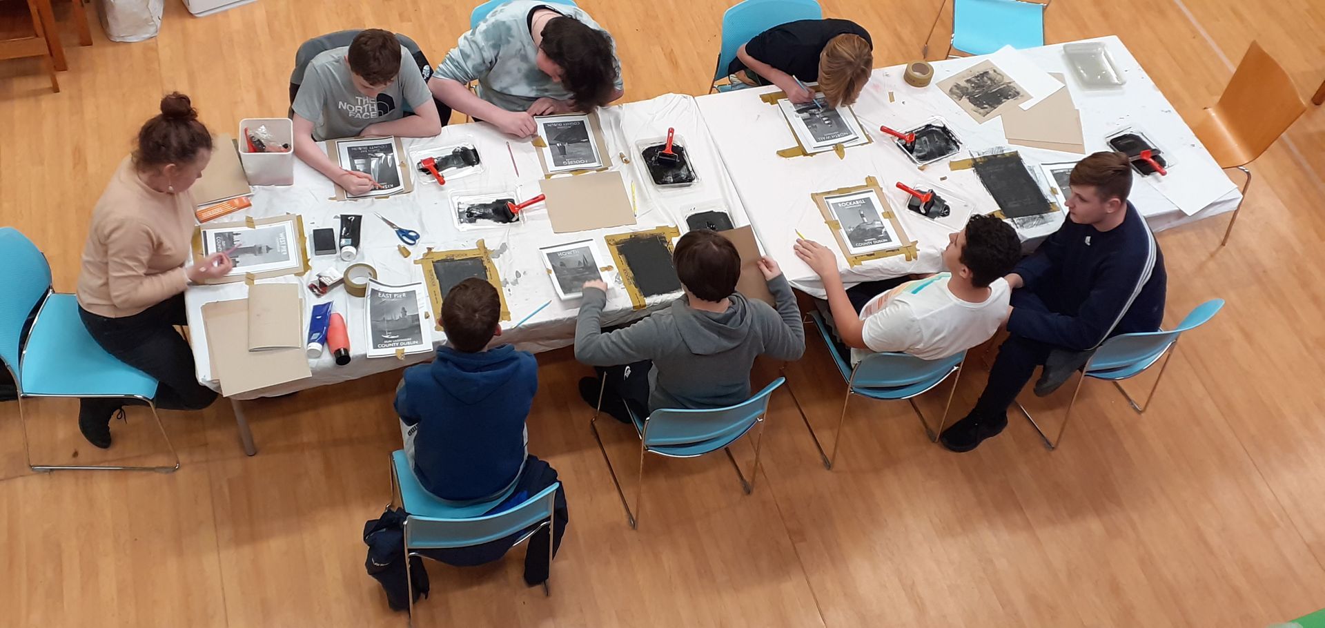 Group of people seated at a long table, working on projects. Interior setting with tools and materials.