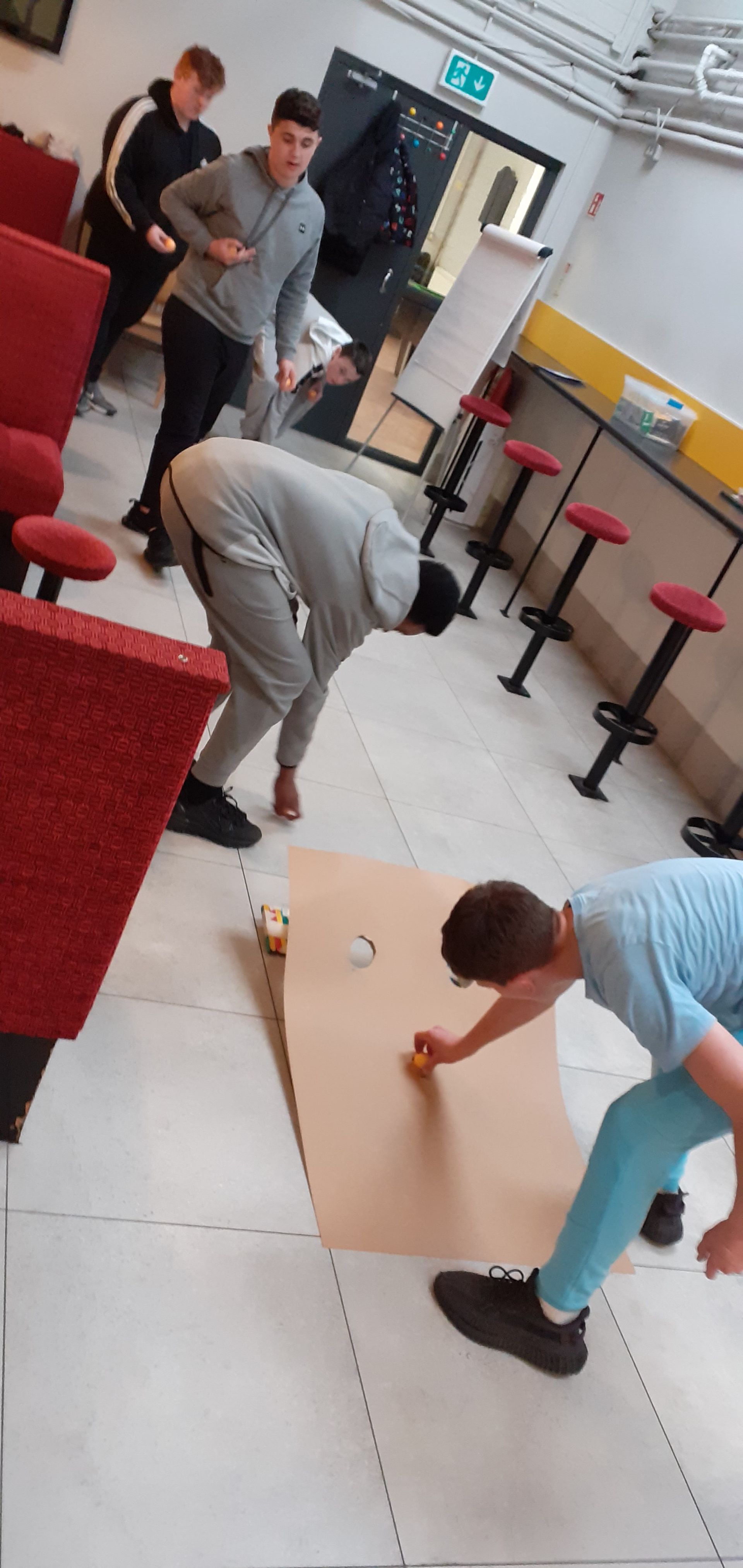 People playing a game with a board on the floor. One person is bending over while another is throwing something.