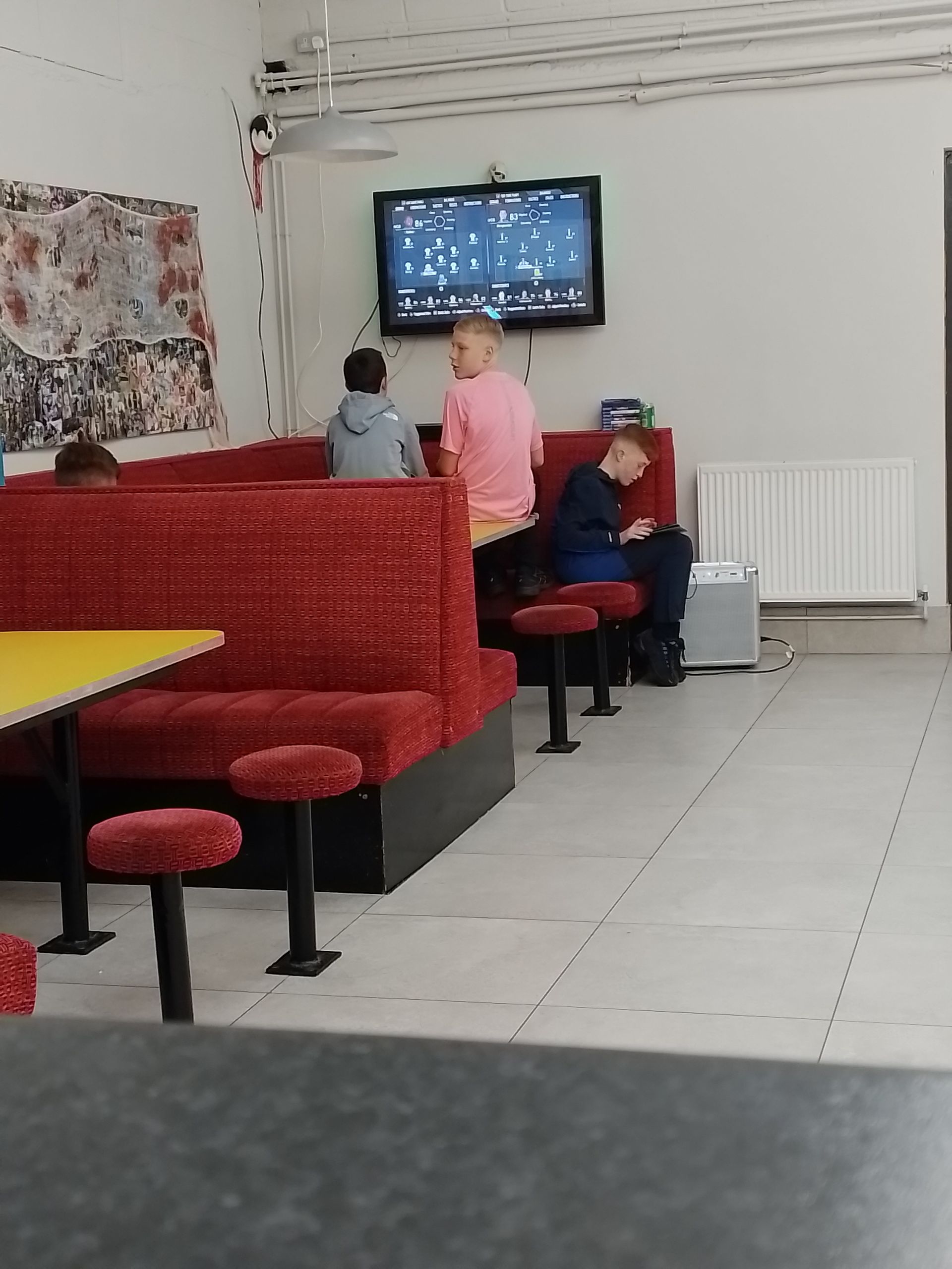 People sit at red booths and stools, watching TV in a room with white walls and a gray floor.