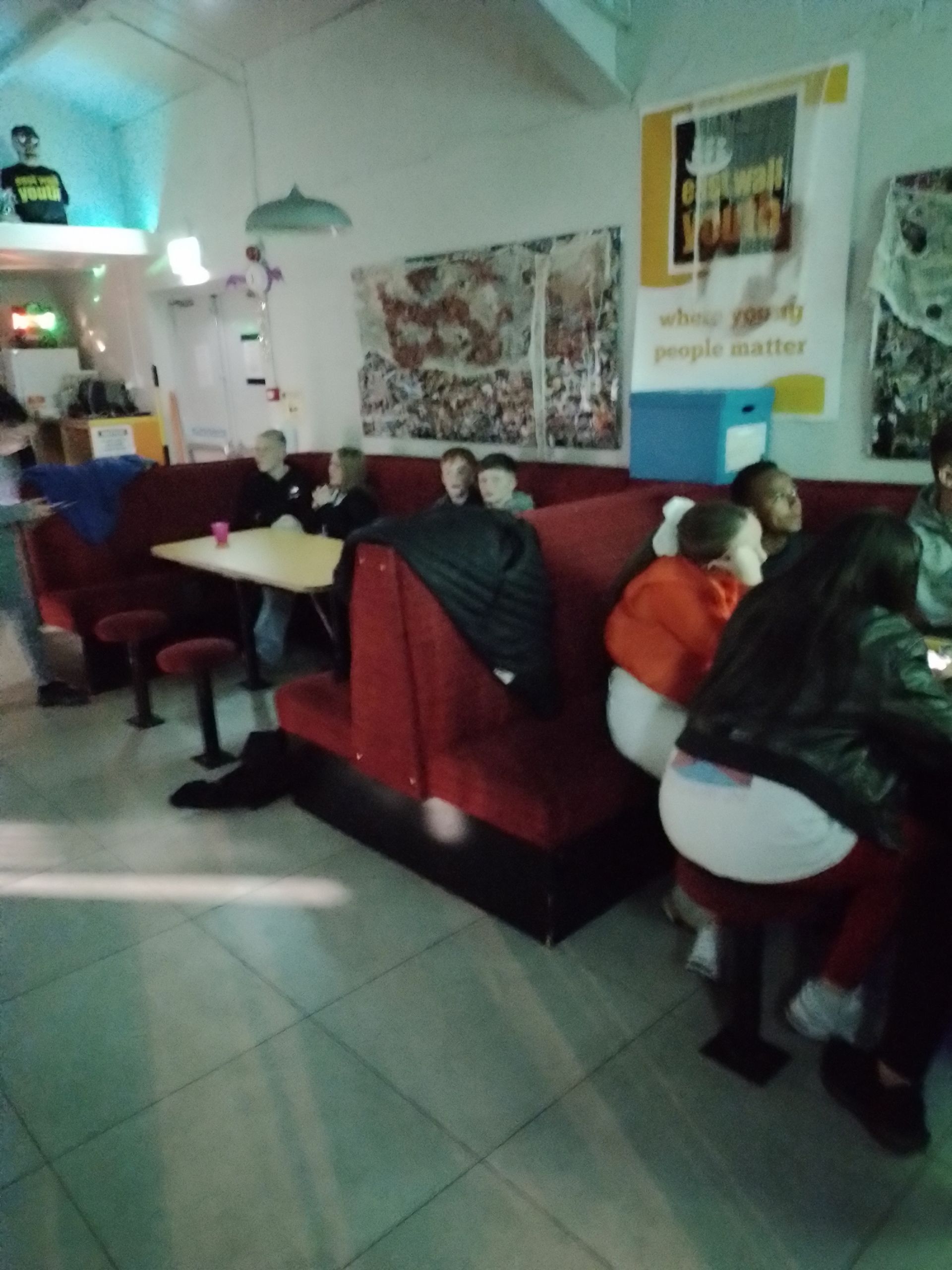 People seated at booths in a diner. Red booths and tables. White walls with posters. Fluorescent lighting.
