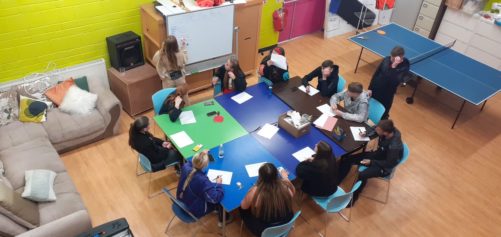 Group of people seated at tables, likely a meeting or workshop. Tables are arranged in a square, with papers on them.
