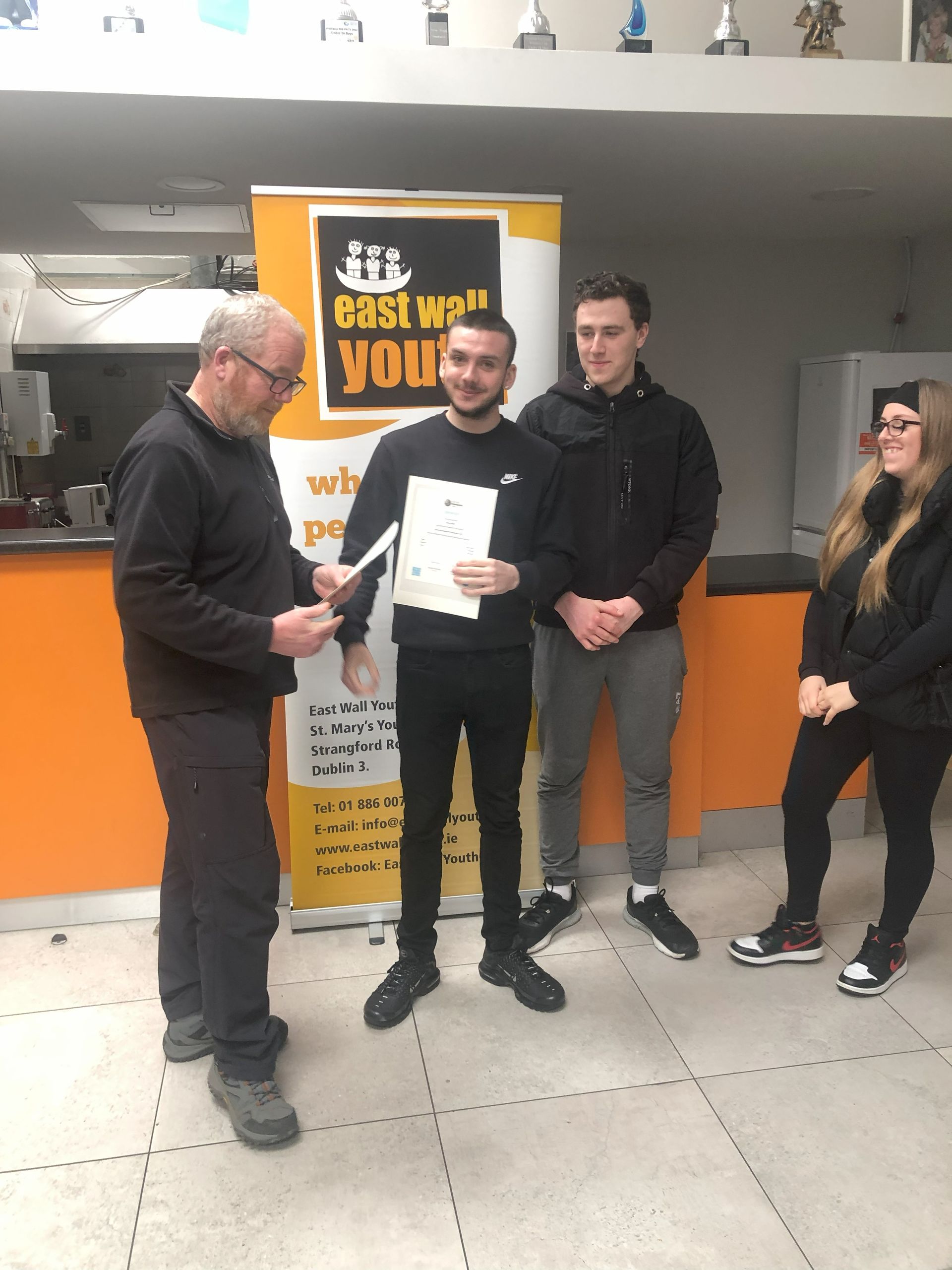 Man presenting a certificate to a young man, two others watch. East Well Youth banner in background.