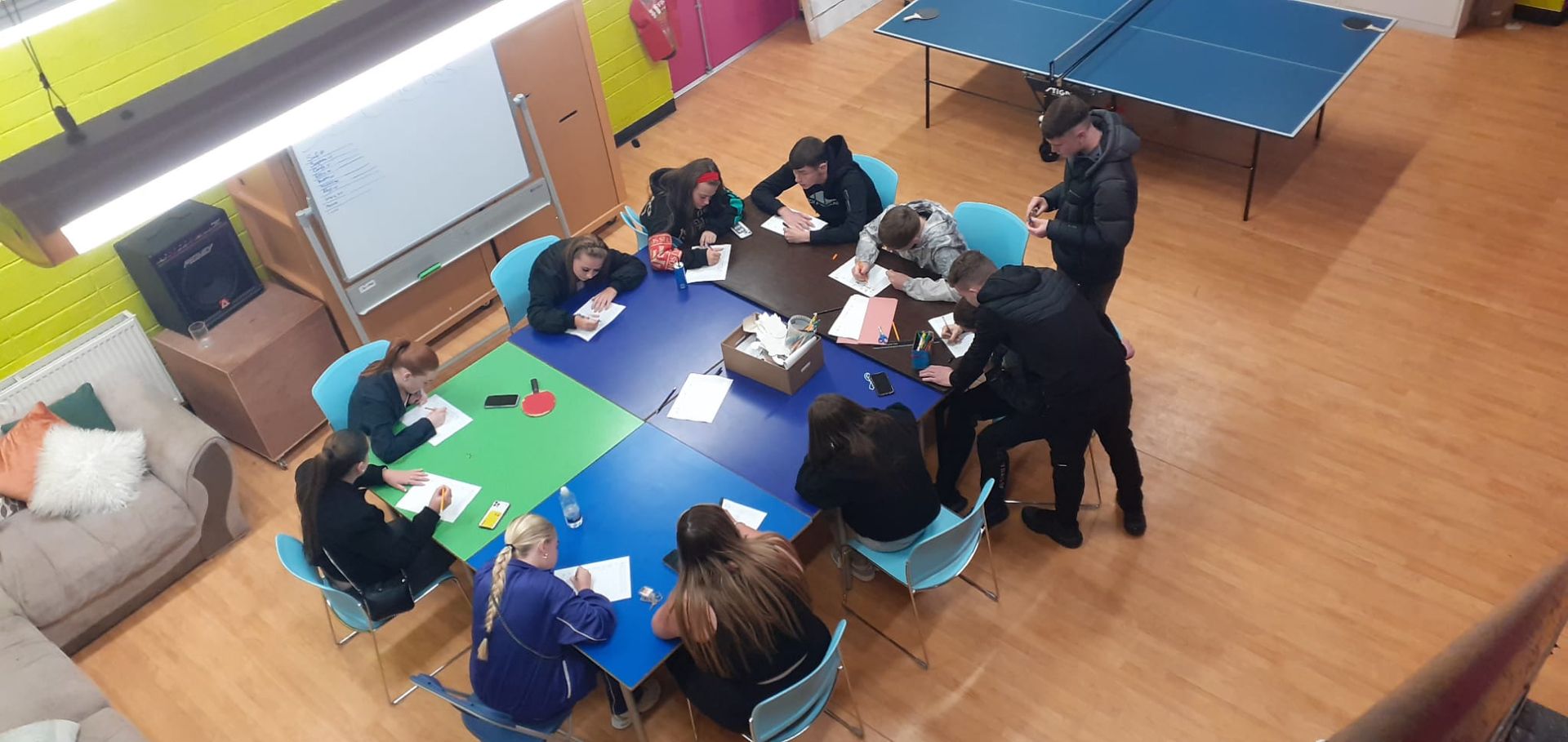 People sitting around a square table, working on papers. Ping pong table in the background. Bright colors and wood floors.