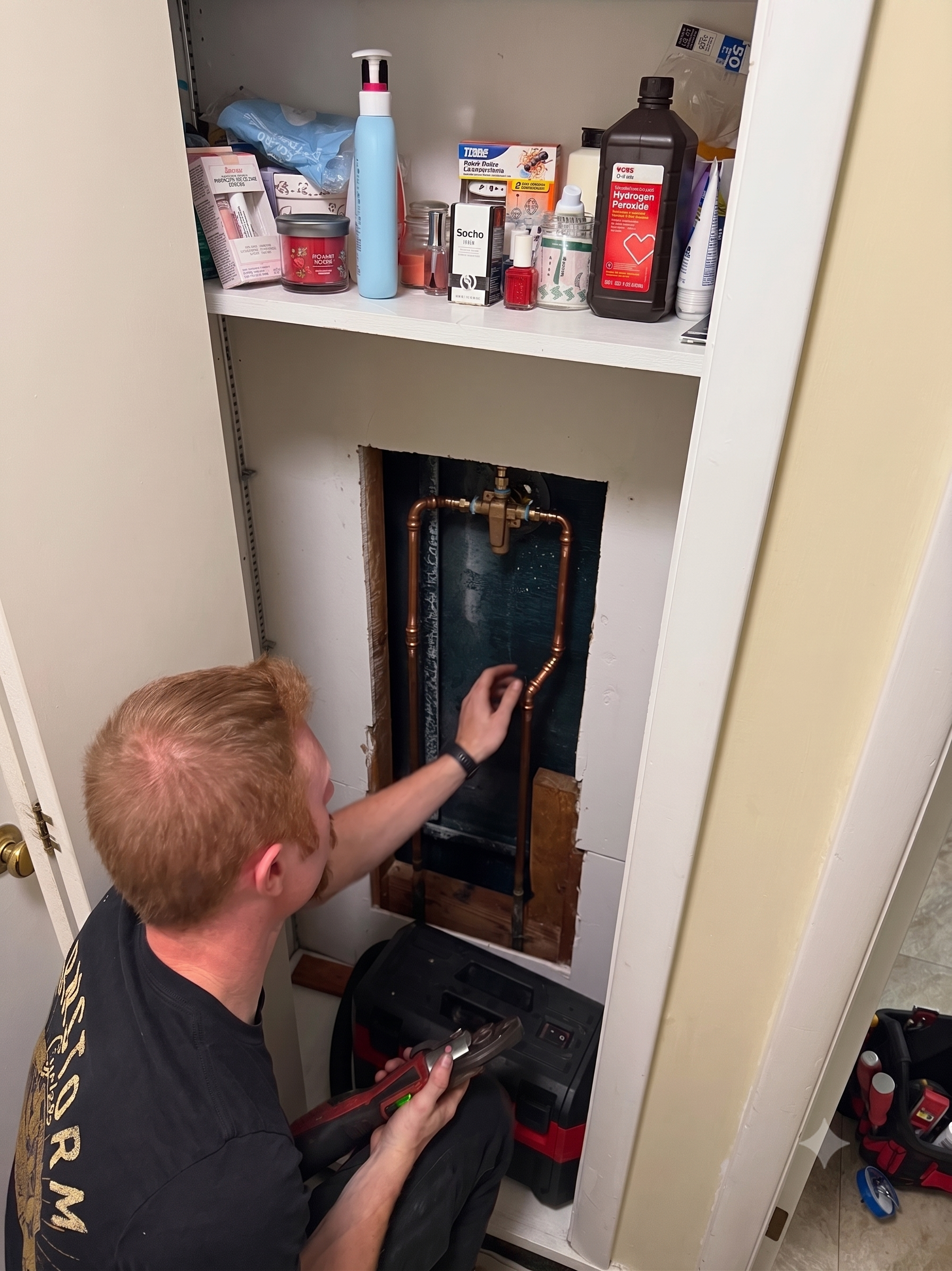Person working inside a wall cavity under a bathroom sink, with tools and supplies on the shelf above