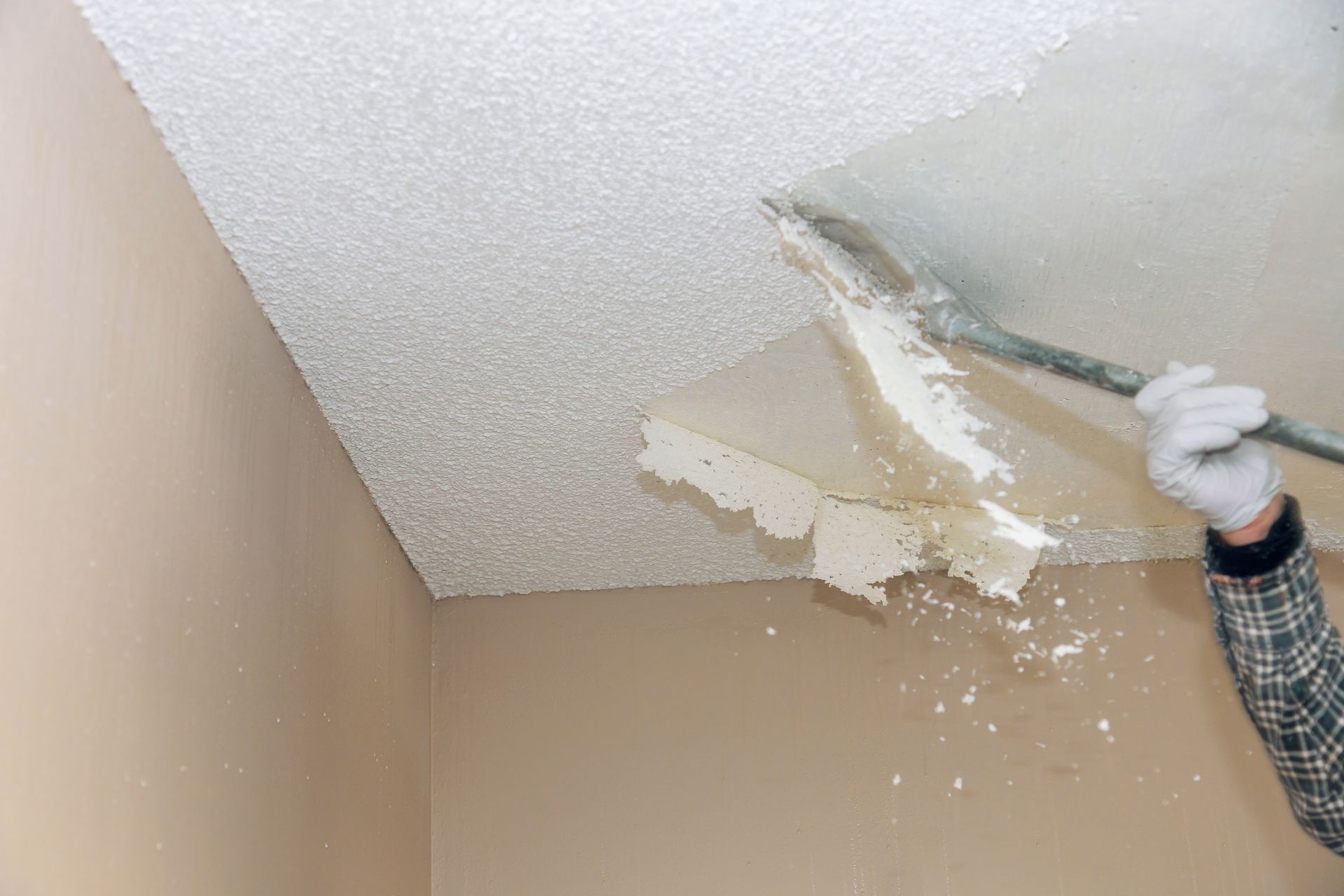 Person scraping peeling ceiling texture with a tool in a beige room, debris falling down