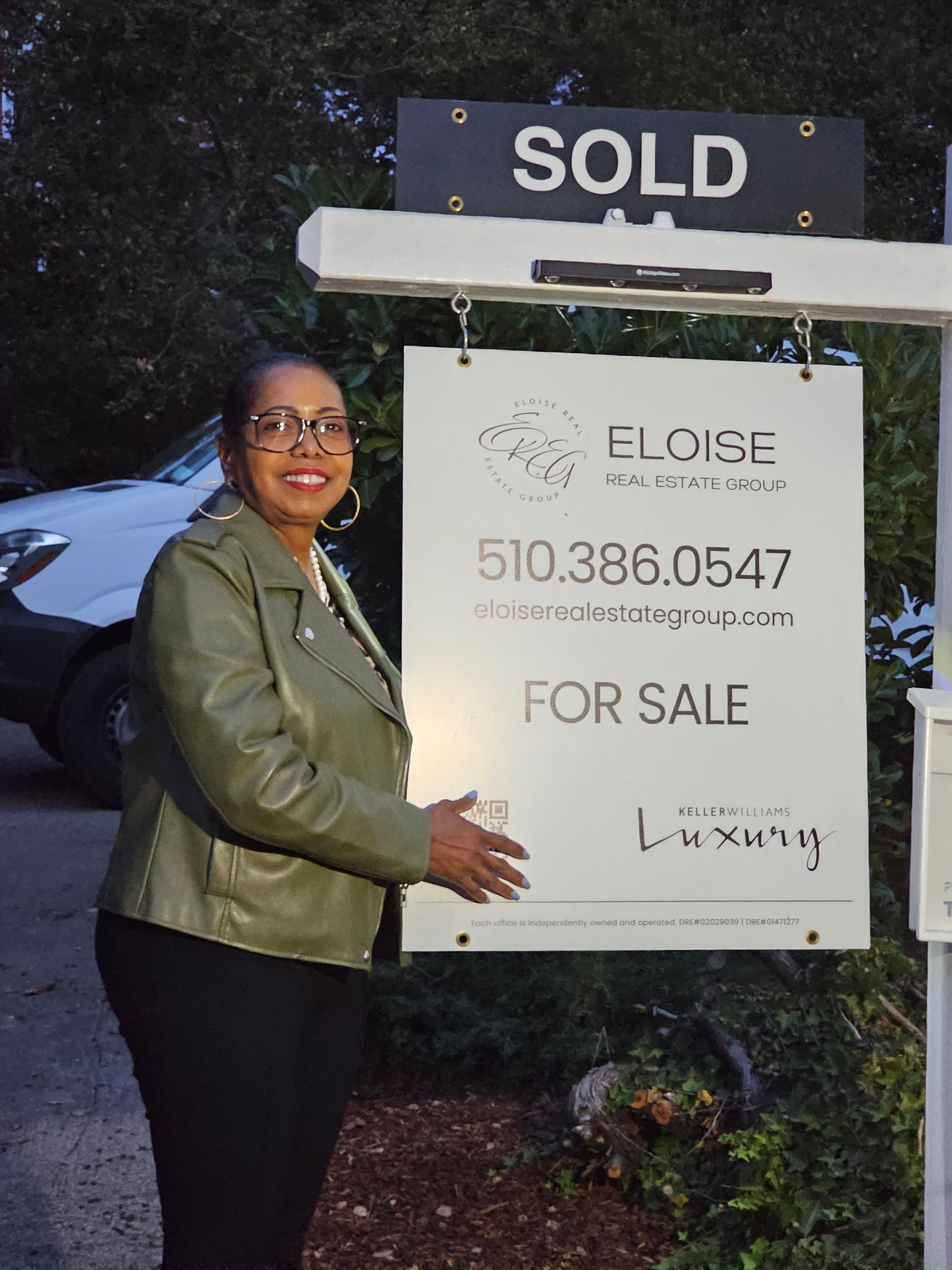 Woman standing beside a real estate sign marked “SOLD” and “FOR SALE” with a house number and phone number.
