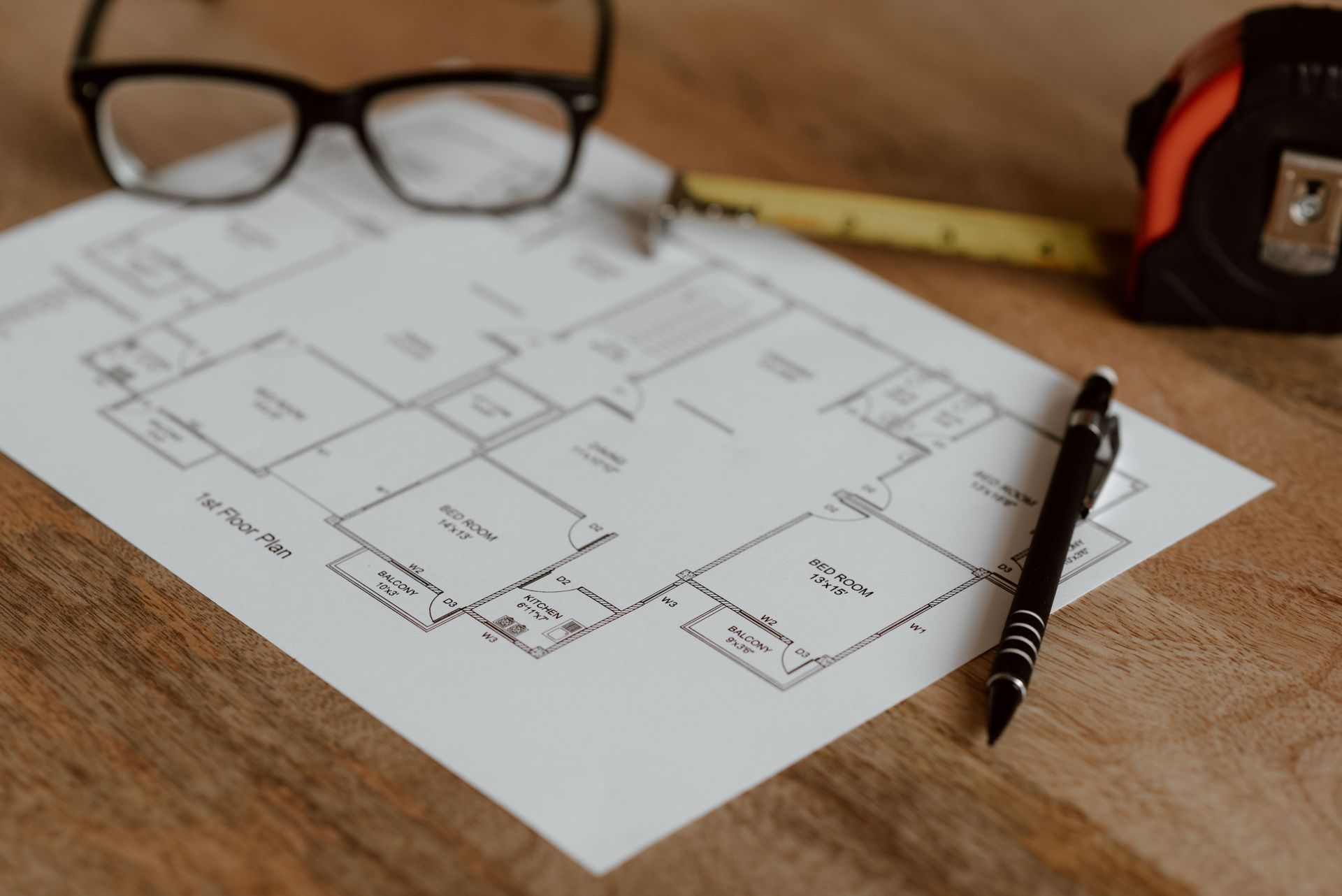 Blueprint on a wooden desk with eyeglasses, a pencil, and a tape measure nearby