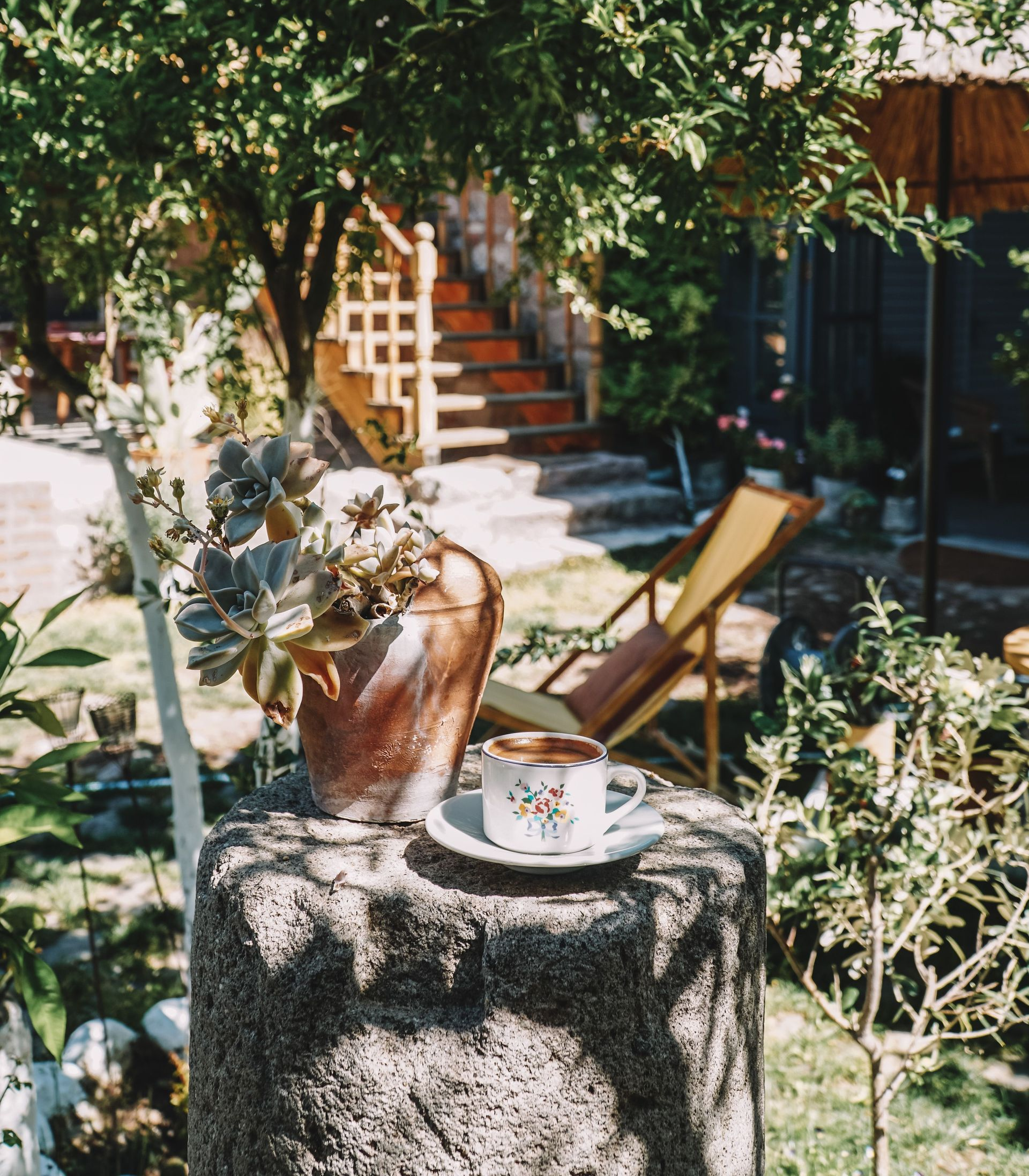 Garden patio with a tree, lounge chair, potted plant, and stone table in sunlight