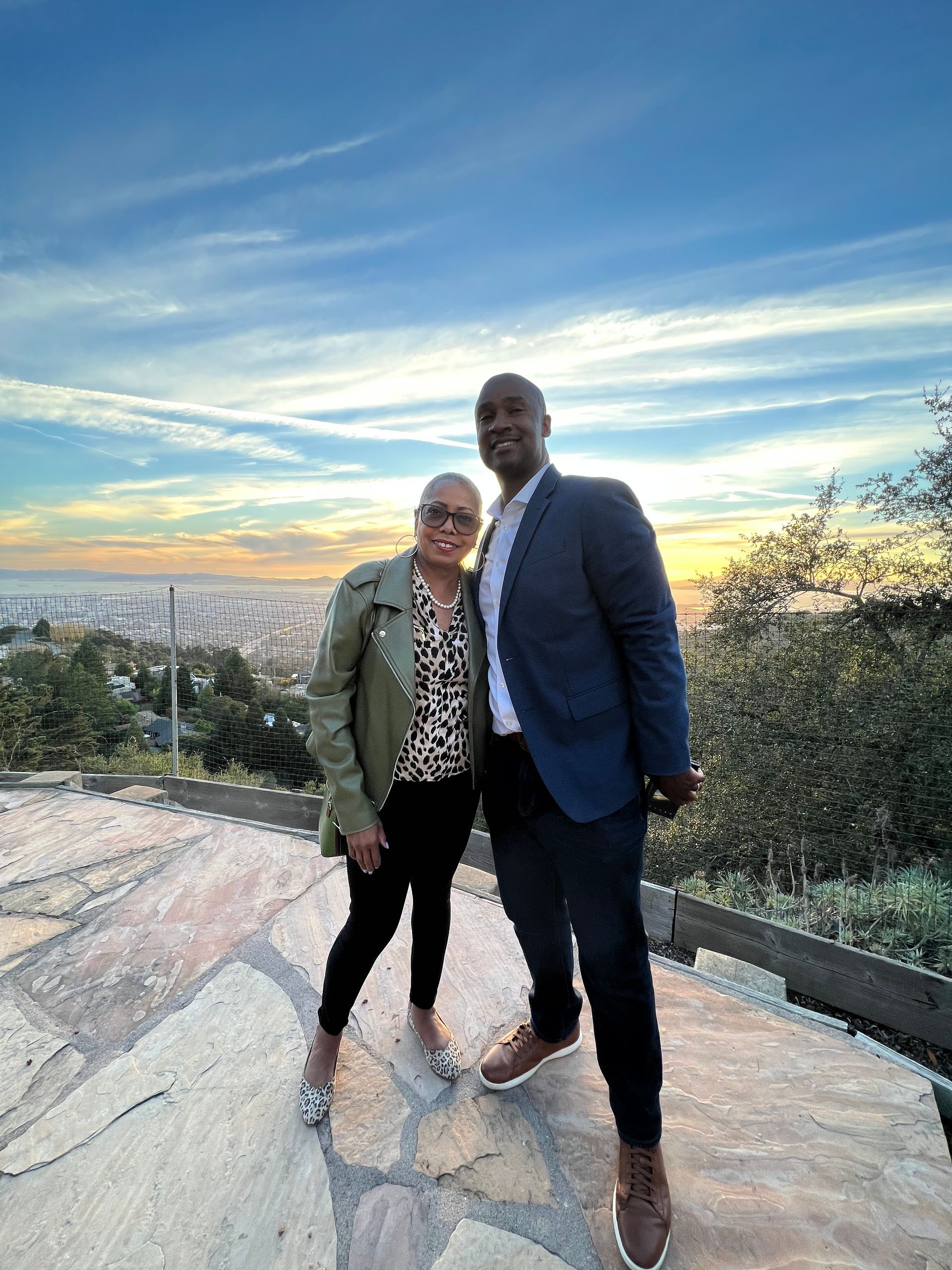 Two people standing on a rocky overlook at sunset, with a city skyline and blue sky behind them