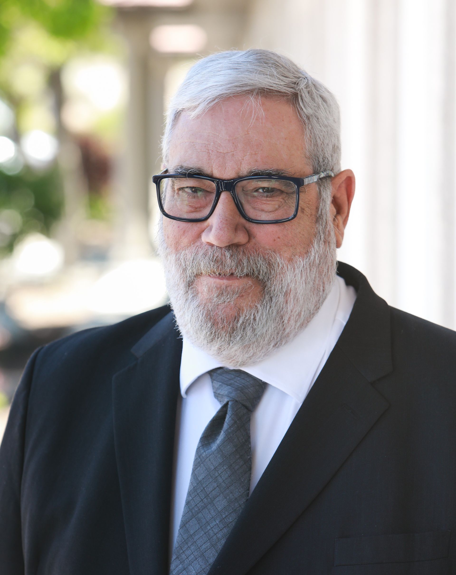 Portrait of a bearded man in glasses and a suit, standing outdoors against a bright background