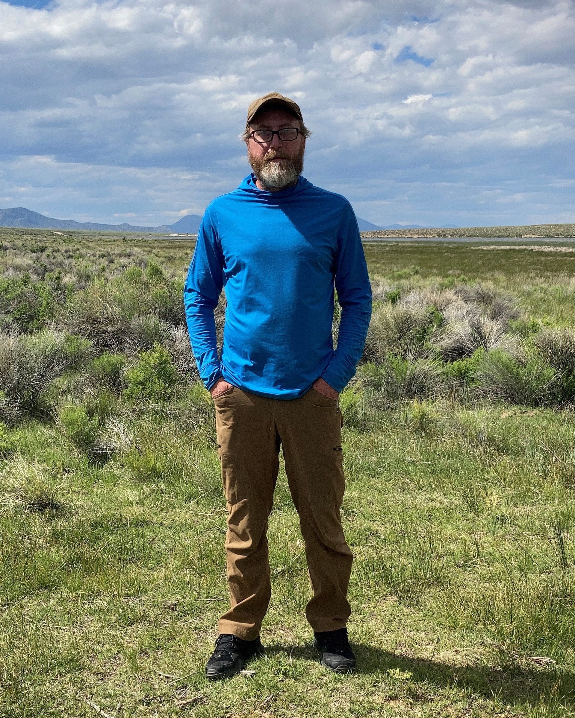 Man standing in a blue jacket and brown pants in a grassy field under a cloudy sky