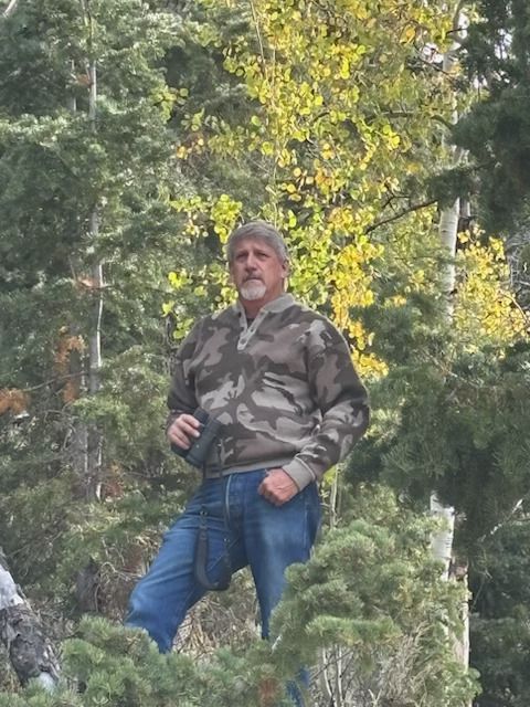 Man in camouflage jacket standing on a rocky forest hillside with trees in the background