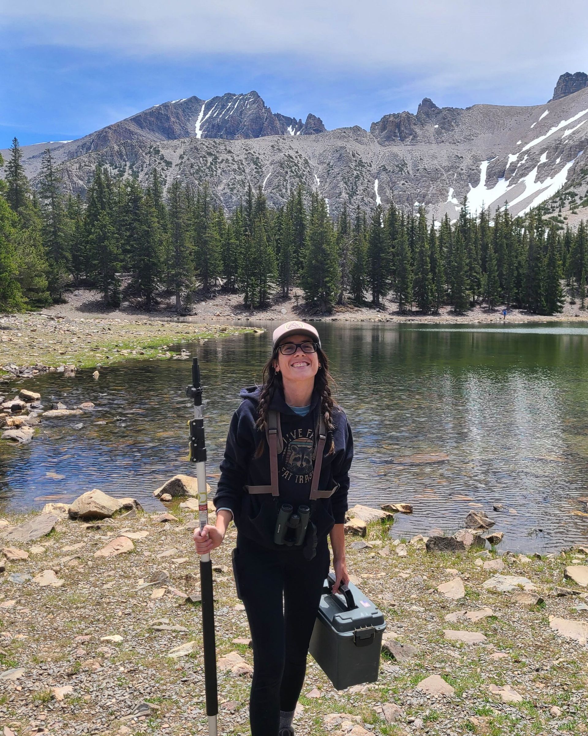 Person standing by a mountain lake holding a hiking pole and cooler, with pine trees and rocky peaks behind.