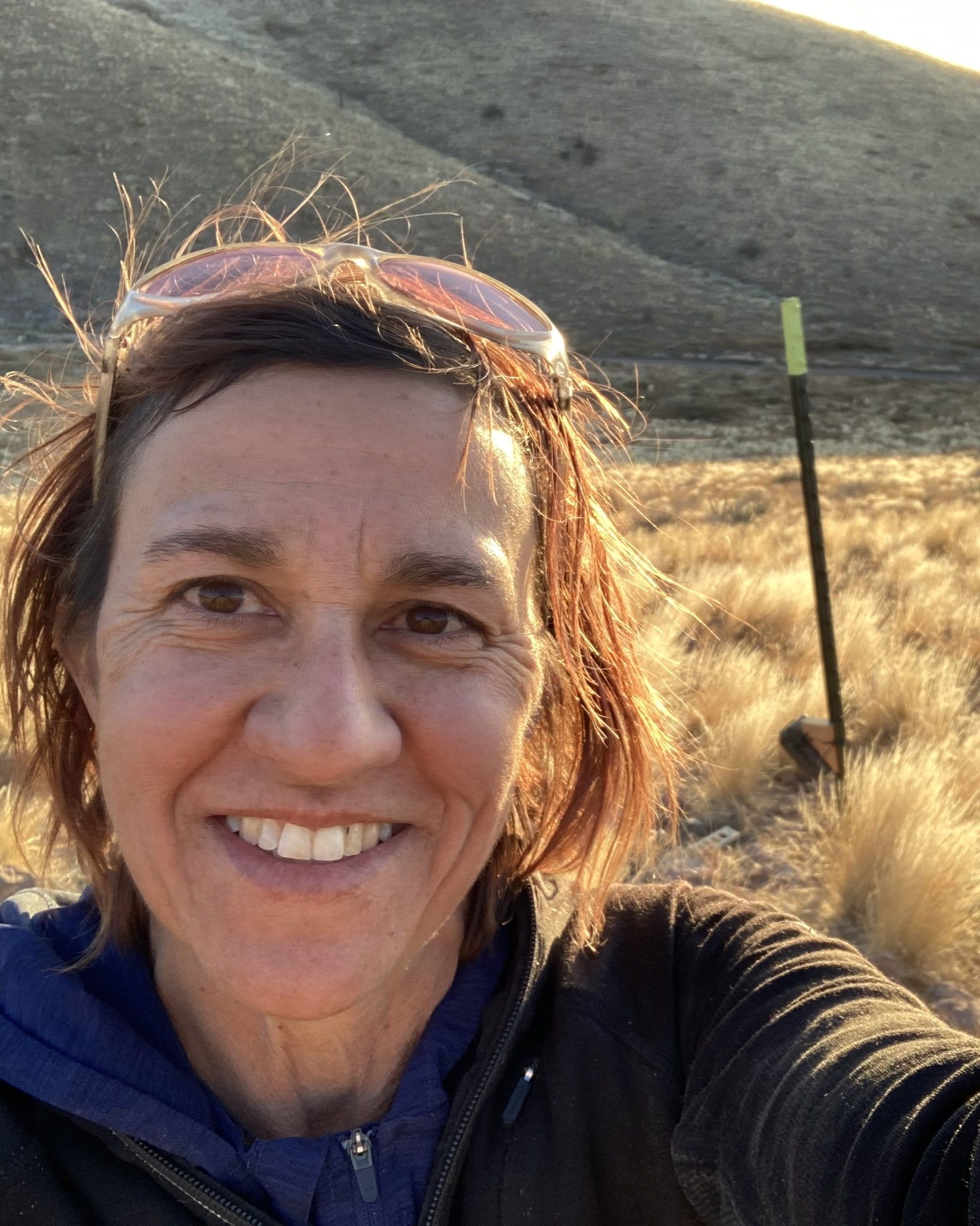Smiling person taking a selfie outdoors on a sunny hillside with dry grass.