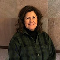 Smiling person in a dark green plaid jacket standing against a stone wall.