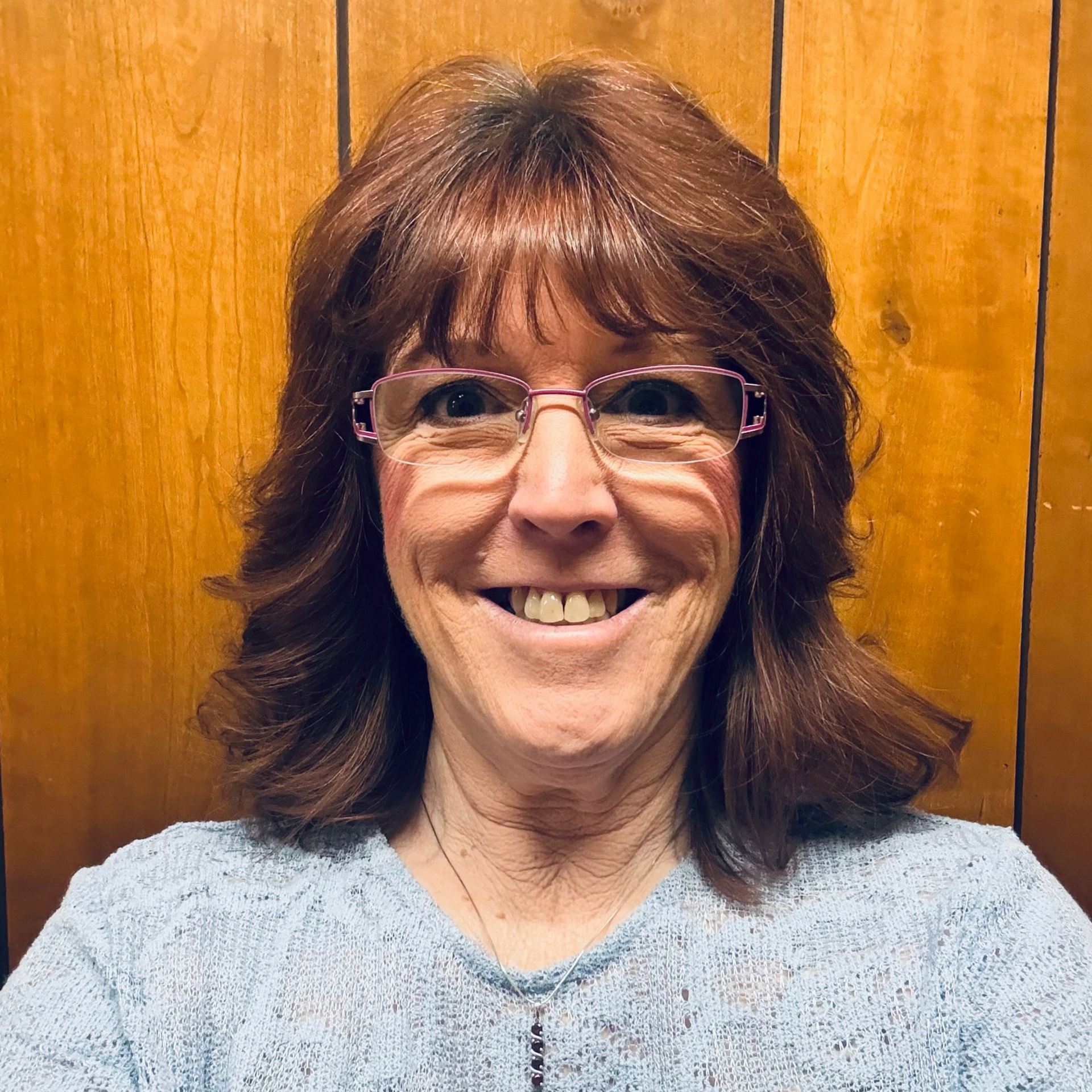 Smiling person with brown hair and glasses in a light blue sweater against a wooden wall