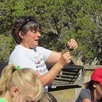 Woman demonstrating a small object outdoors while children watch nearby