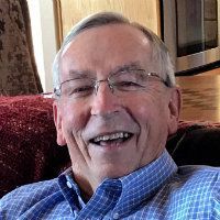 Smiling man with glasses sitting indoors on a couch, wearing a blue checkered shirt