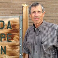 Man standing beside a wooden sign against a brick wall, wearing a plaid shirt