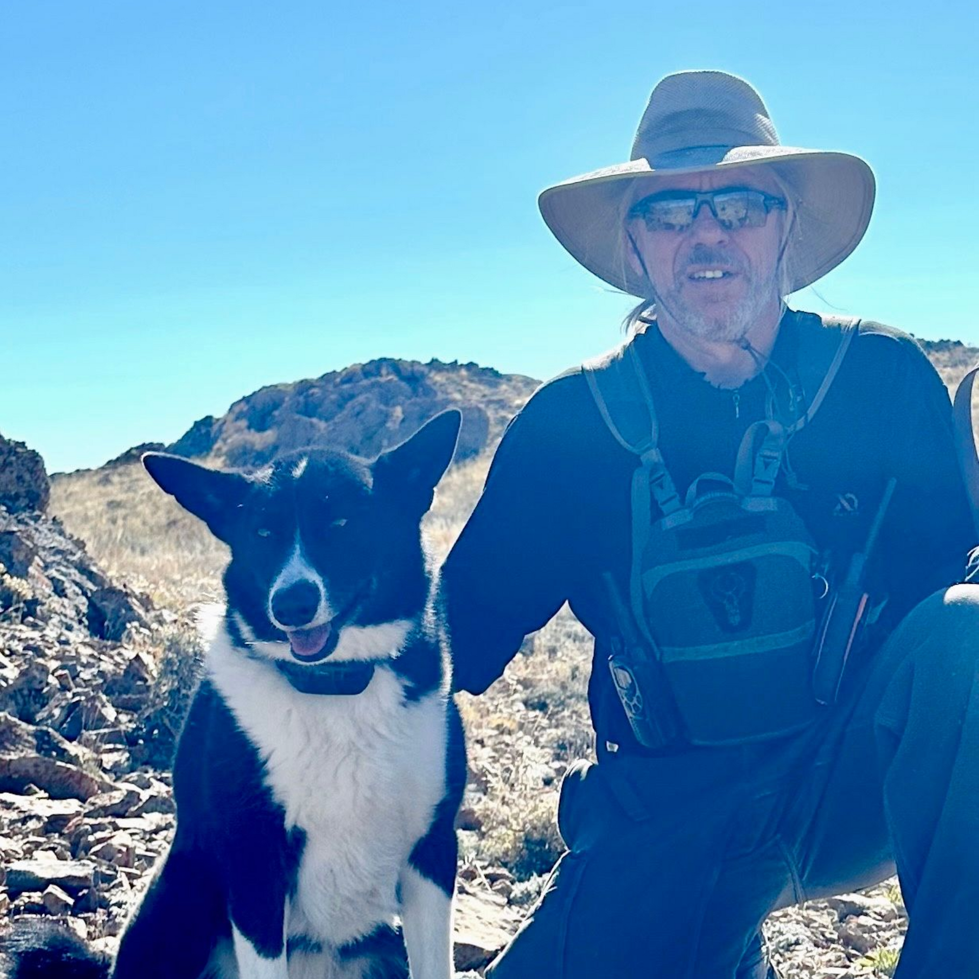 Man in a hat posing with a black-and-white dog on a rocky mountain trail under a blue sky